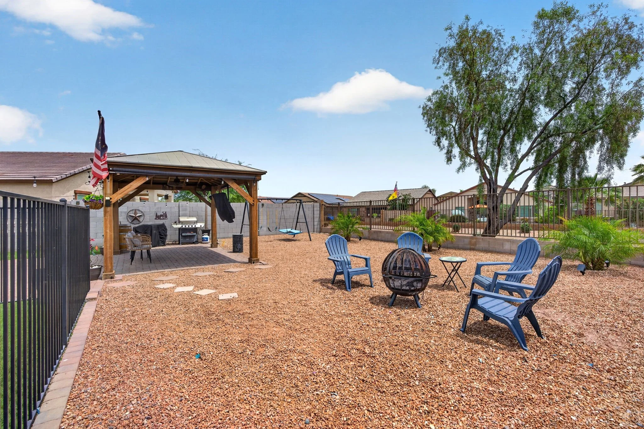 A backyard patio area with a gravel surface, blue chairs, a fire pit, a wooden pergola, a swing set, and a cinder block wall. There are houses with solar panels and trees in the background, under a bright blue sky with scattered clouds.