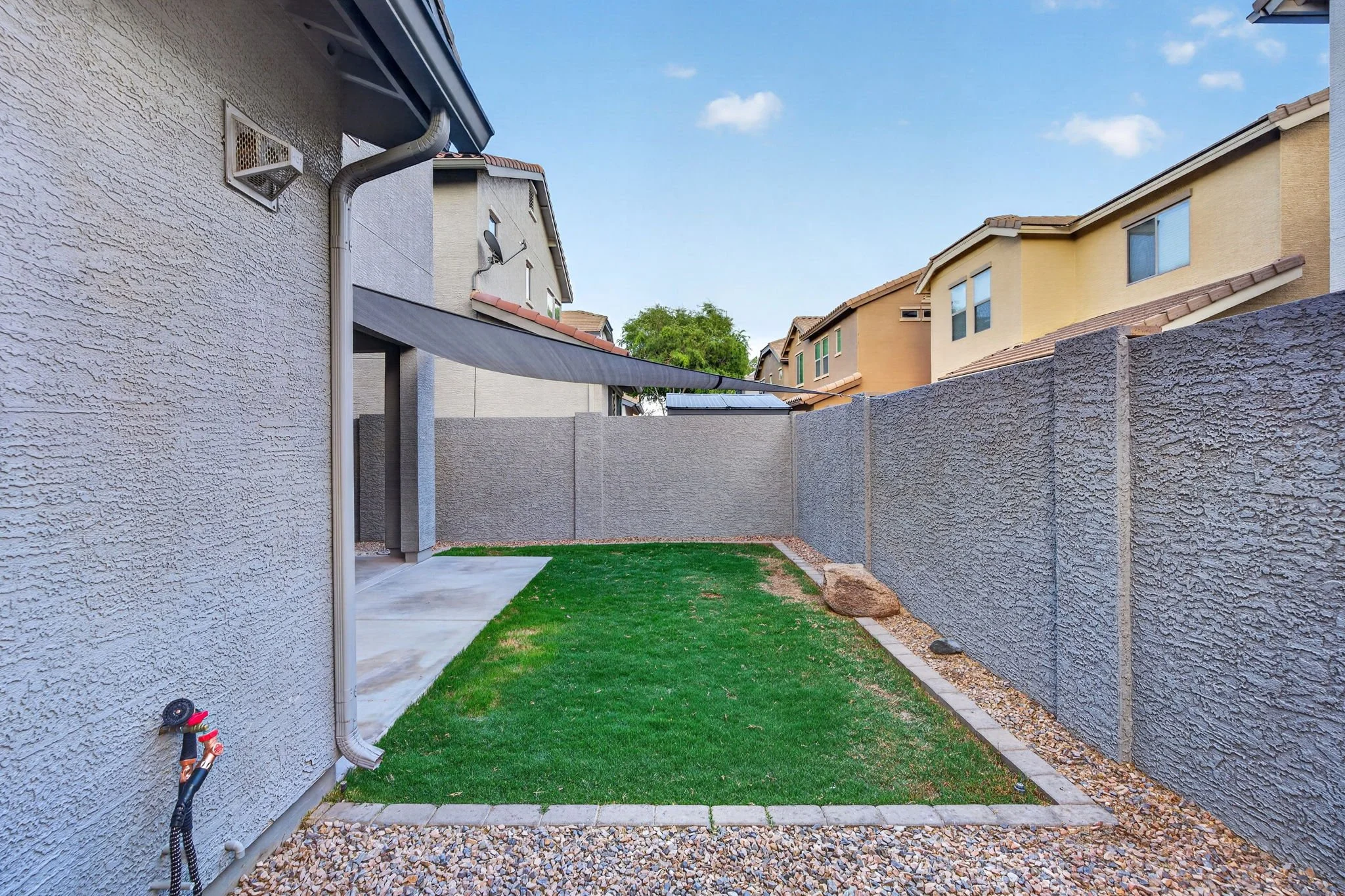 Backyard with grass, a concrete patio, a gravel border, and a tall gray stucco fence. Houses with beige and gray exteriors and tiled roofs are visible beyond the fence. A downspout and a hose are attached to the house on the left.