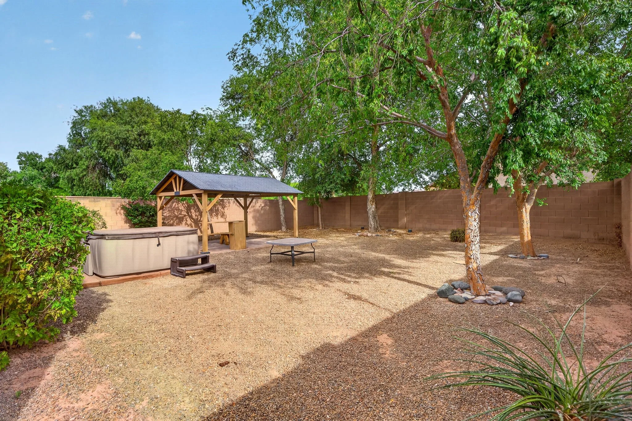 A backyard with a gravel ground, multiple trees, a brick wall, a wooden pergola with a metal roof, and outdoor furniture including a hot tub, a small table, and a bench.