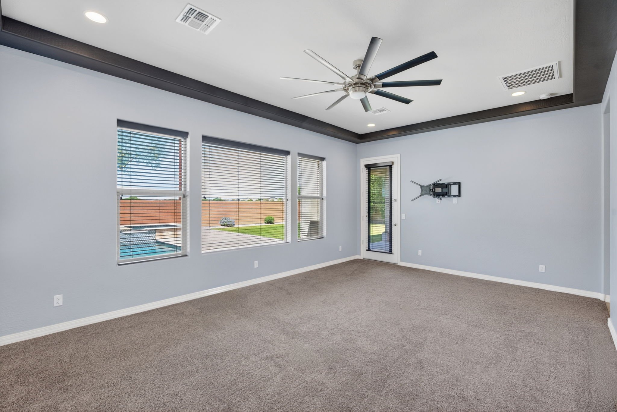 Empty living room with gray carpet, white walls, large windows with blinds, and a ceiling fan.