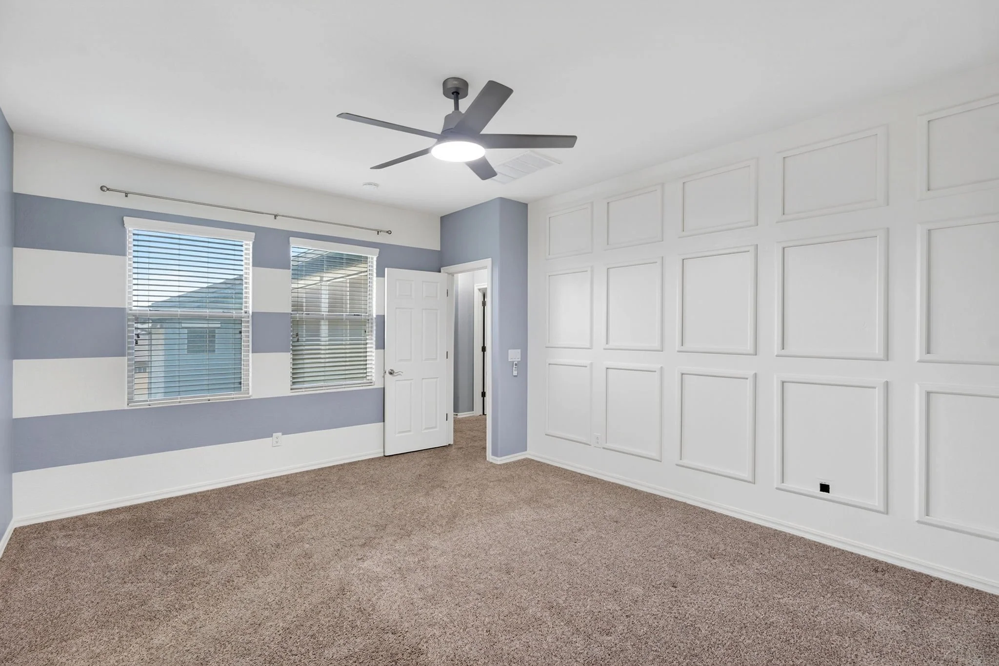 Empty room with blue and white painted walls, large white paneled wall, windows with blinds, ceiling fan with light, carpeted floor, and open door.