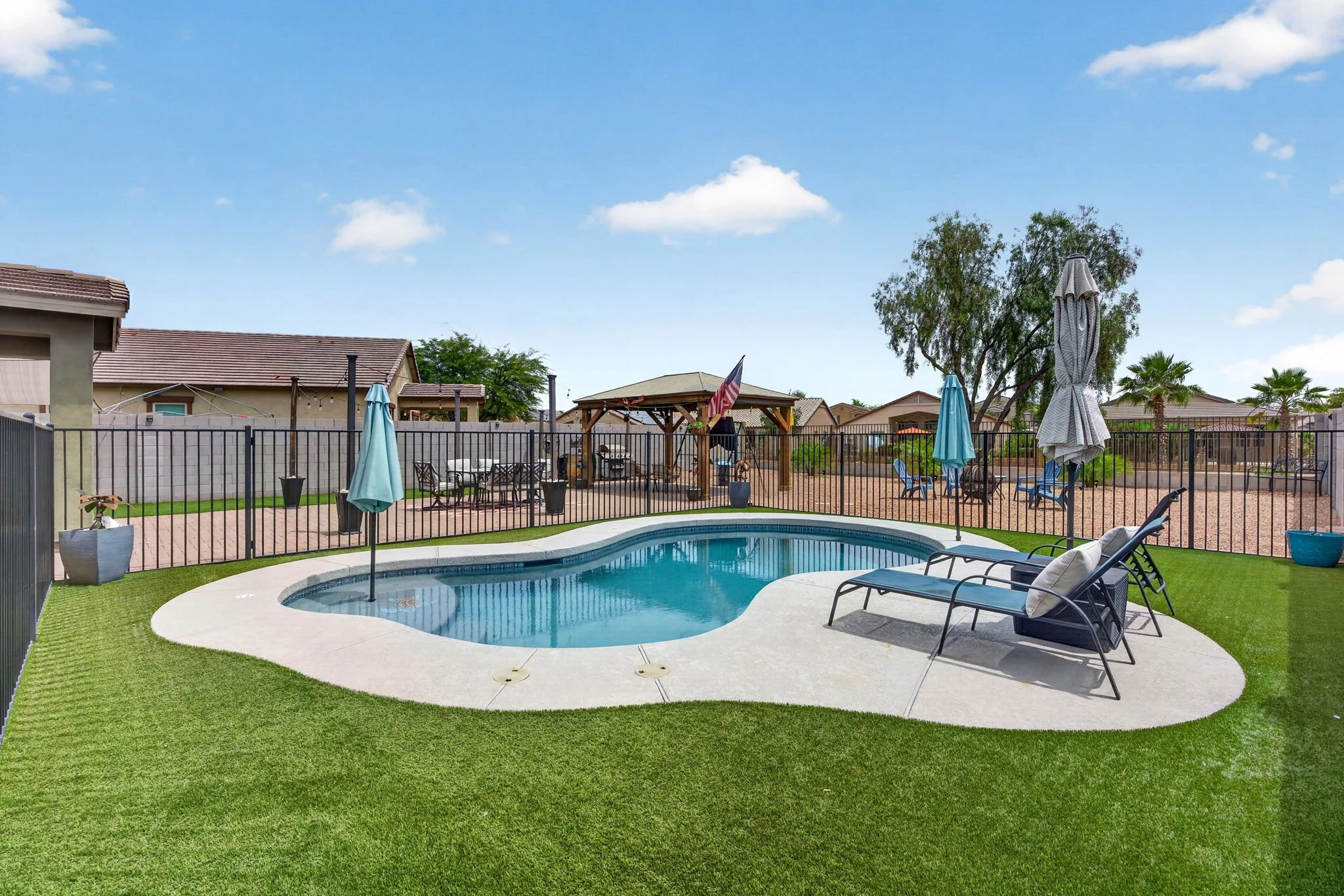Backyard with swimming pool, sun loungers, umbrellas, and a fenced yard with houses and trees in the background.