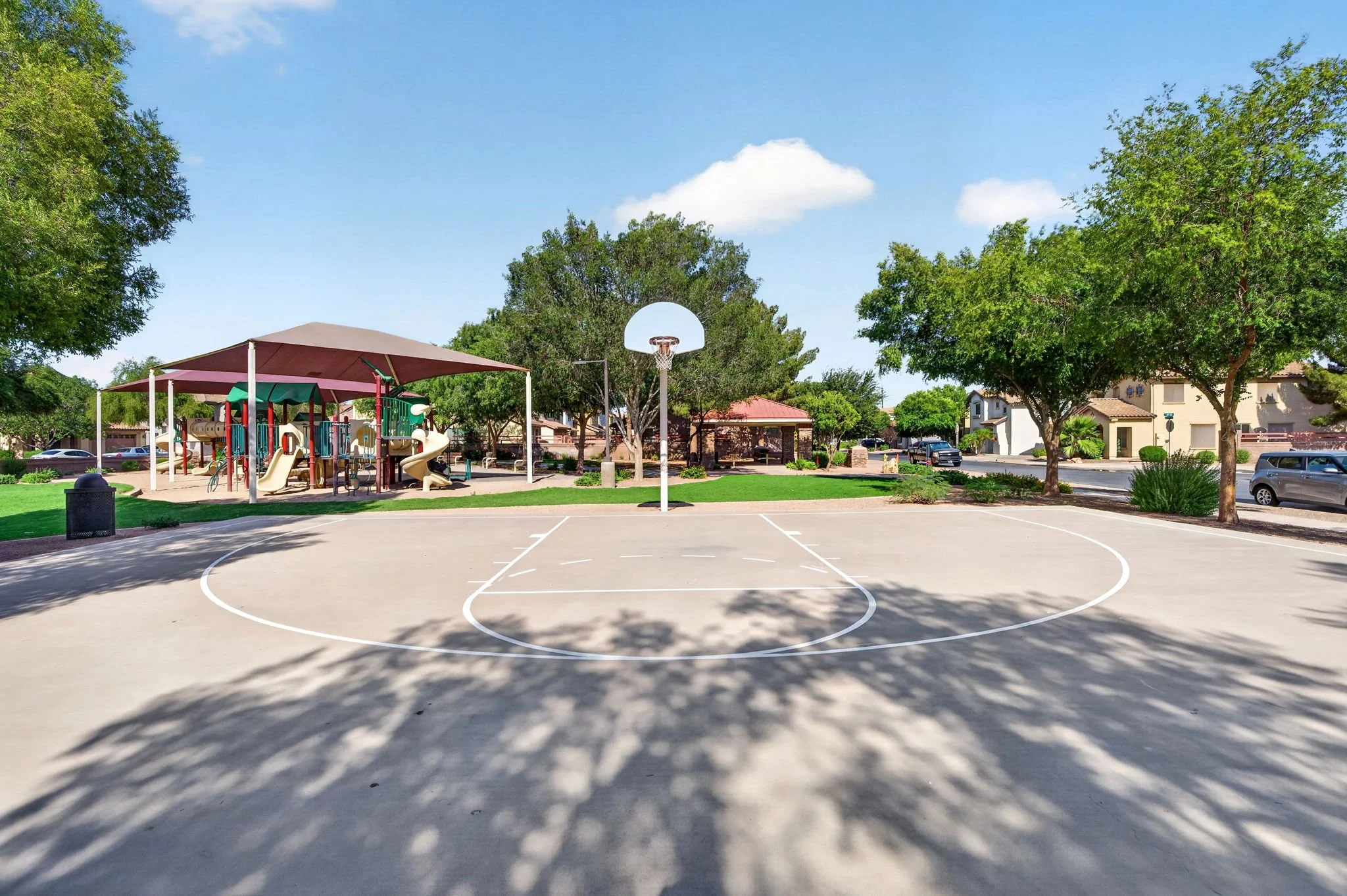 An empty outdoor basketball court with a hoop, surrounded by trees, houses, and parked cars under a blue sky with clouds.