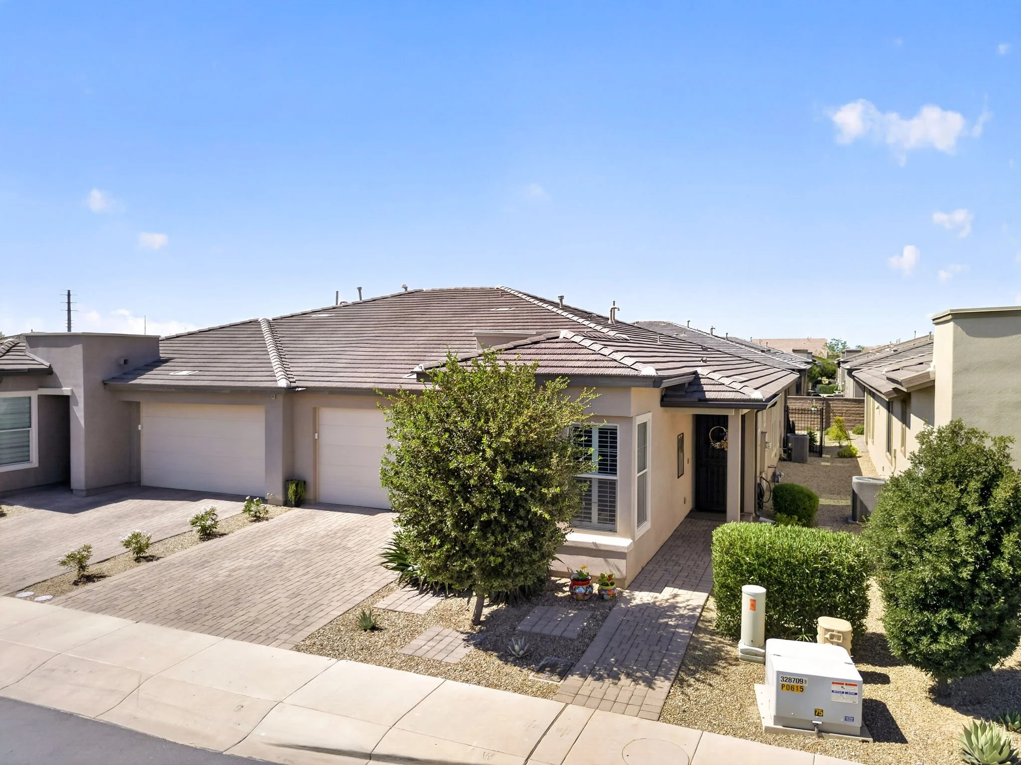 View of a suburban neighborhood with houses featuring light-colored walls and gray tile roofs. The front yards are landscaped with gravel, small bushes, and a few potted plants. There are green trees and a paved walkway in the foreground, with a clea
