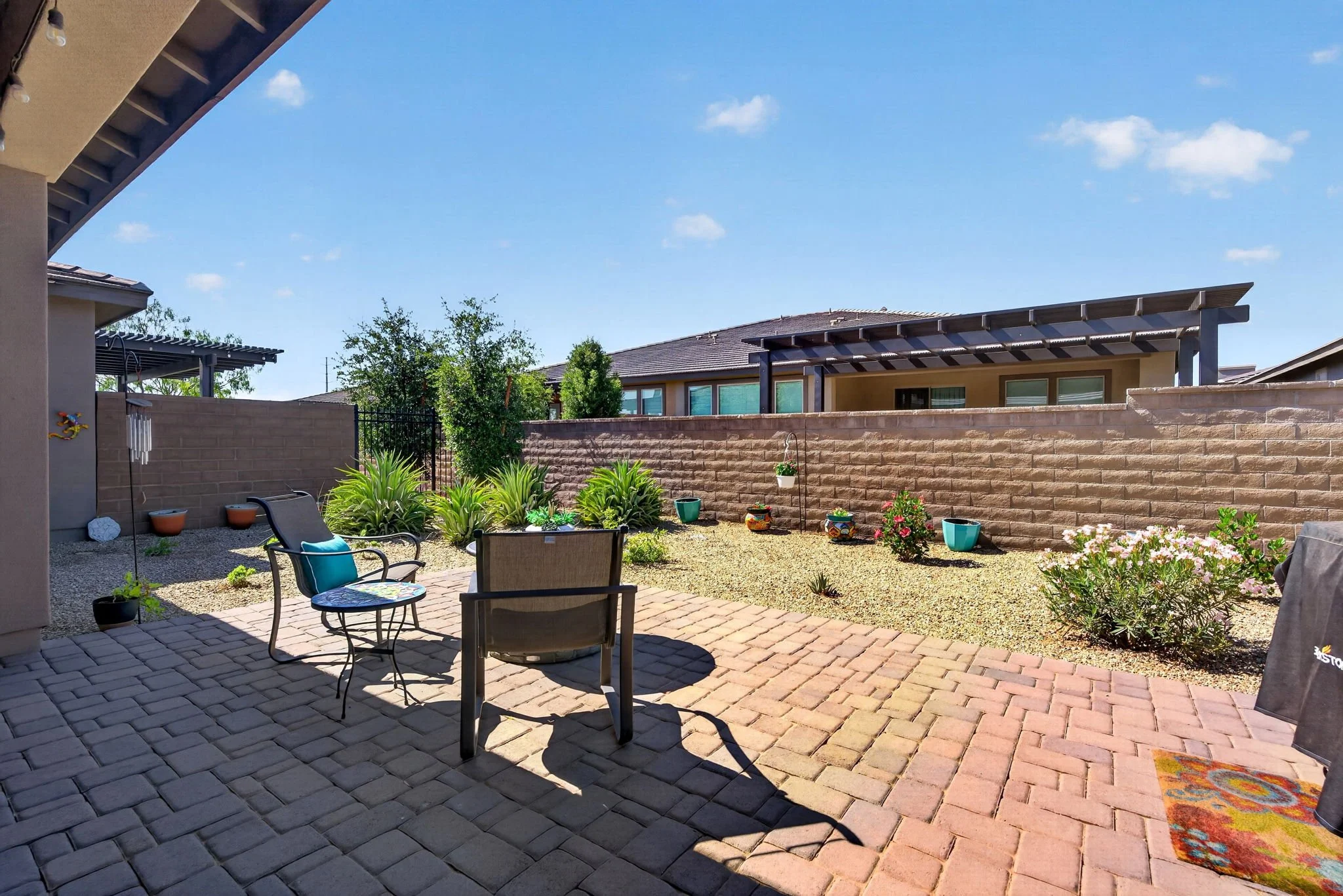 A backyard patio with chair and table, garden plants, potted flowers, a brick and gravel ground, neighboring houses with modern flat roofs, and a bright blue sky with a few clouds.