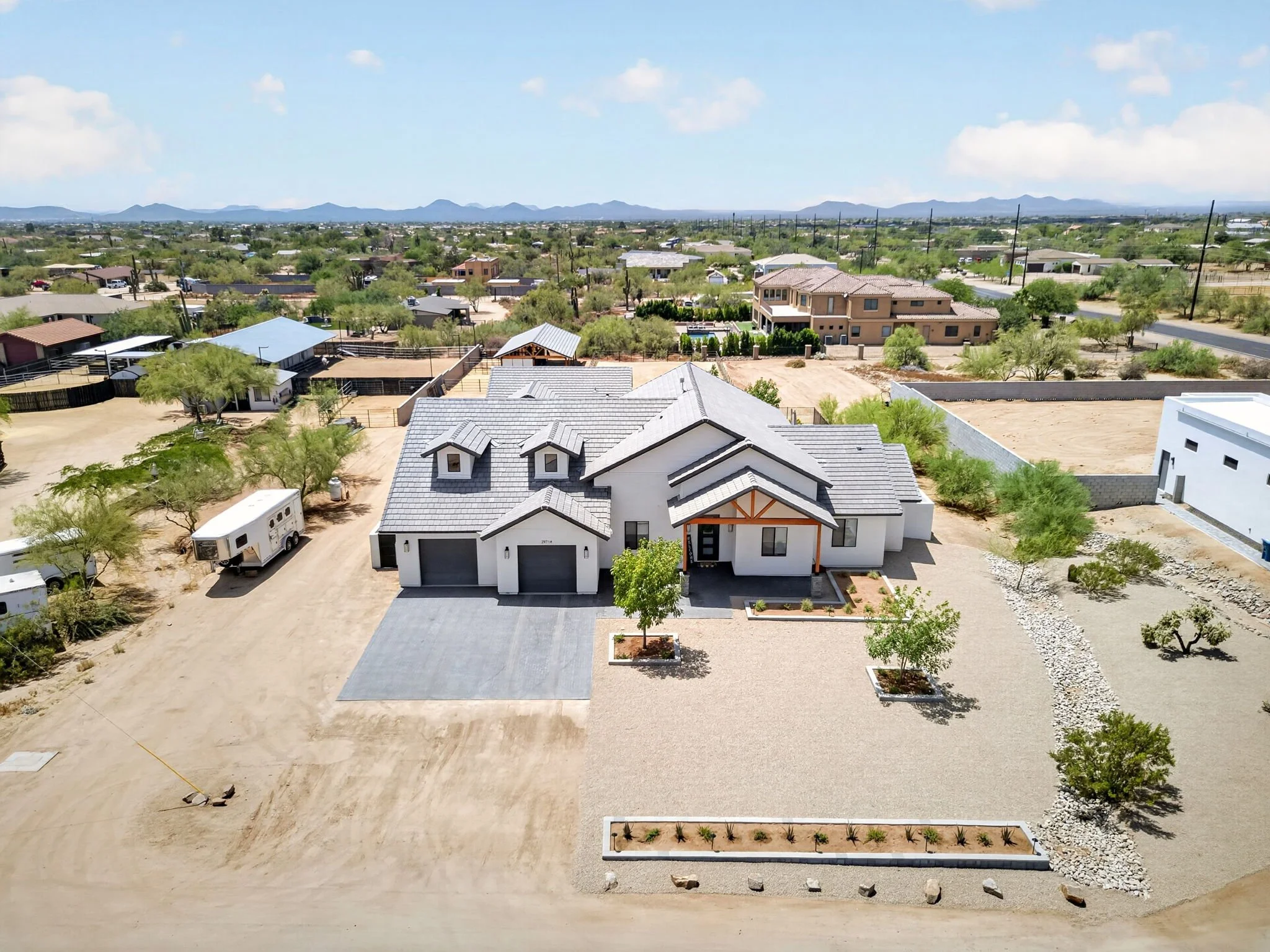Aerial view of a suburban neighborhood in a desert landscape showing houses, trees, and open yards, with mountains in the background under a clear blue sky.