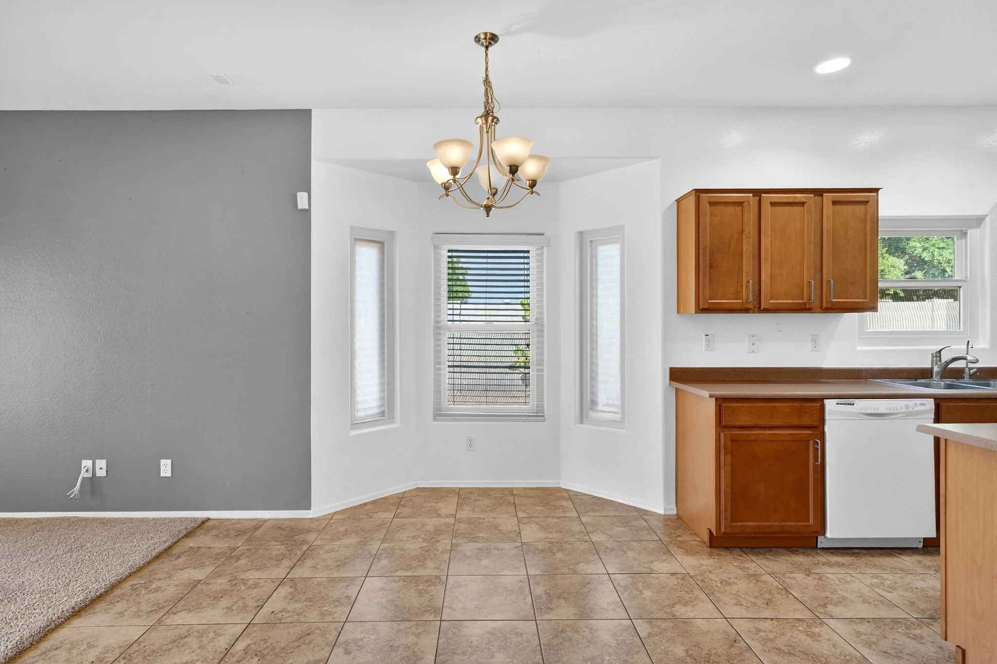 Interior of a kitchen with beige tiled floor, wooden cabinets, a chandelier, and three windows with white blinds.