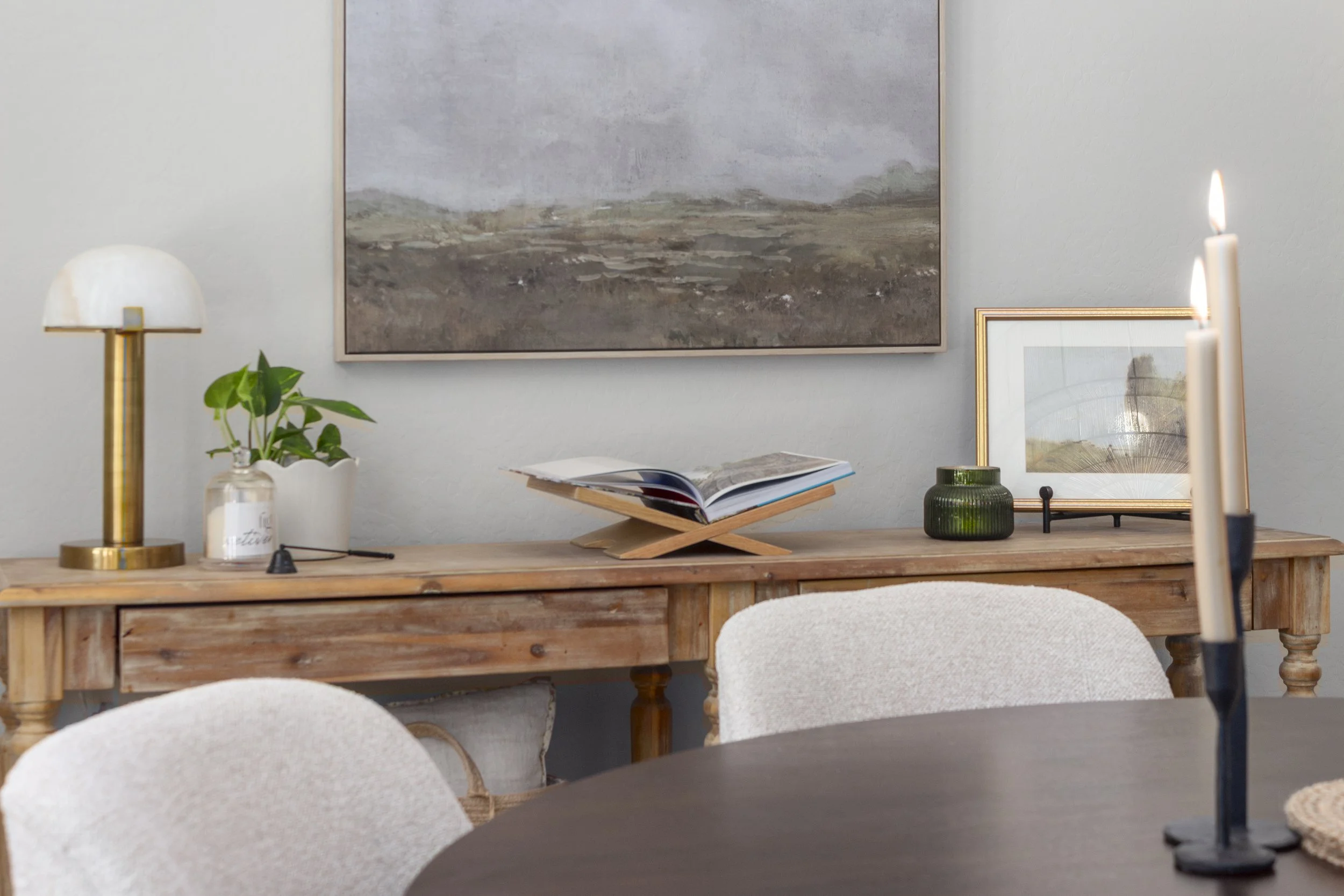 A wooden sideboard with a potted plant, a book, framed pictures, and a table lamp on it, set against a wall with a large landscape painting.