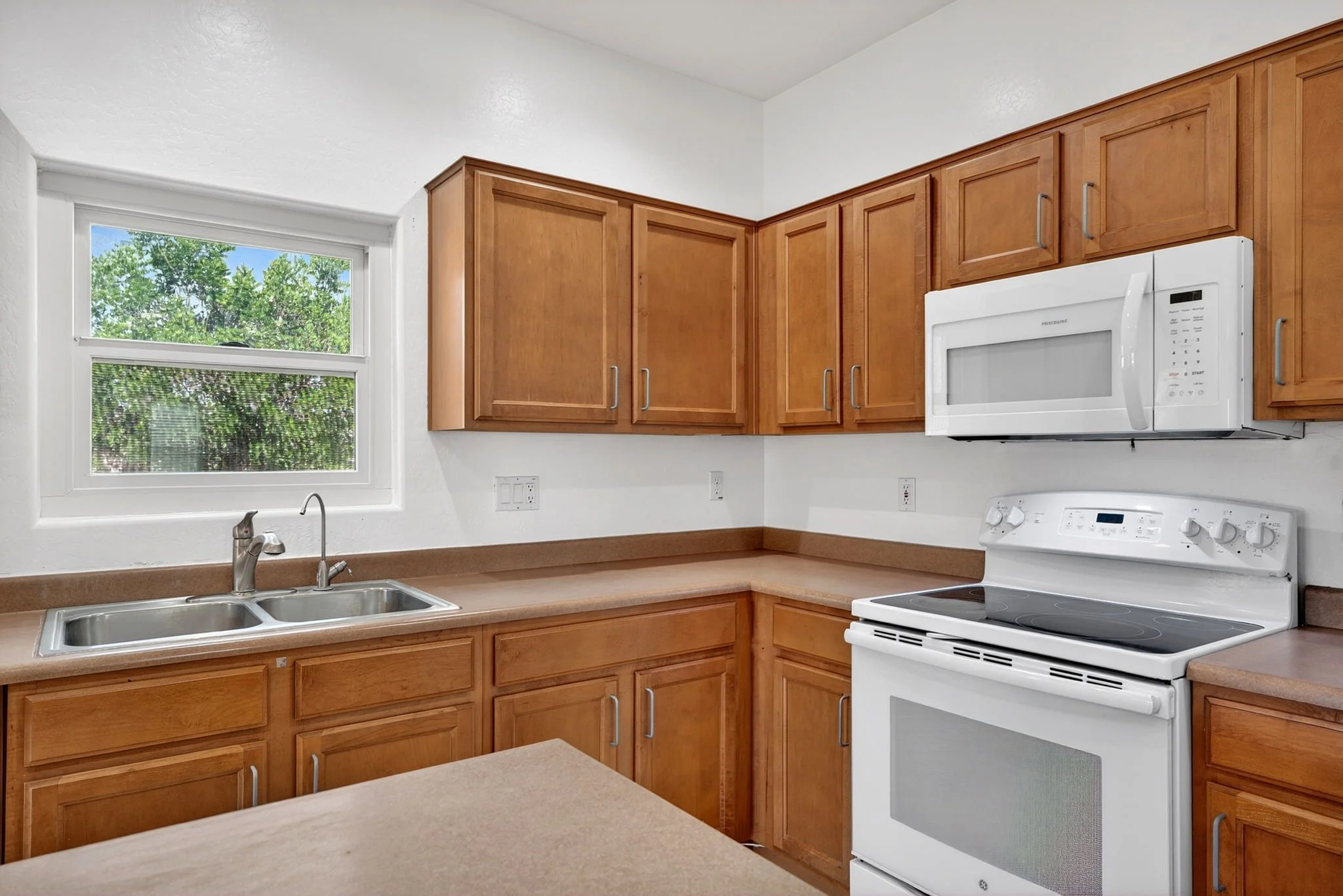 Kitchen with wooden cabinets, a window showing trees outside, a white microwave above an electric stove, and a double sink under the window.