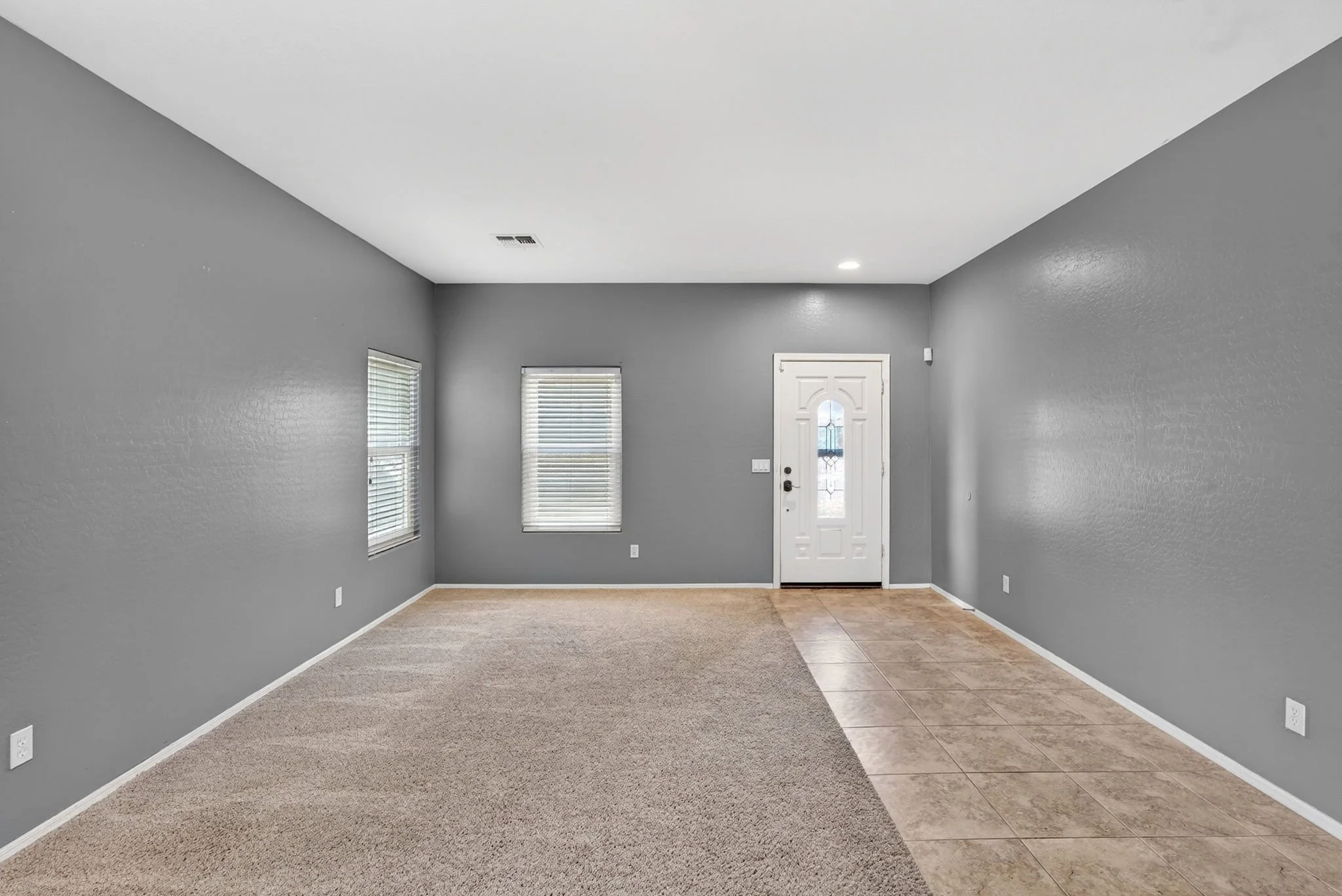 Empty living room with gray walls, beige carpet, tiled entry area, and front door with glass panel.