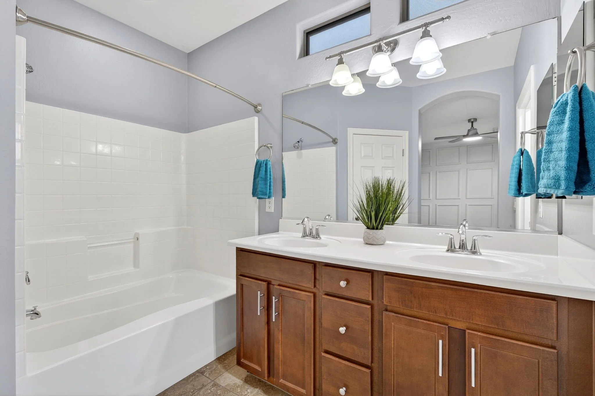 Bathroom with a double sink vanity, wooden cabinets, large mirror, light fixture with four bulbs, potted plant, blue towels, white tiled shower area, and a ceiling fan.