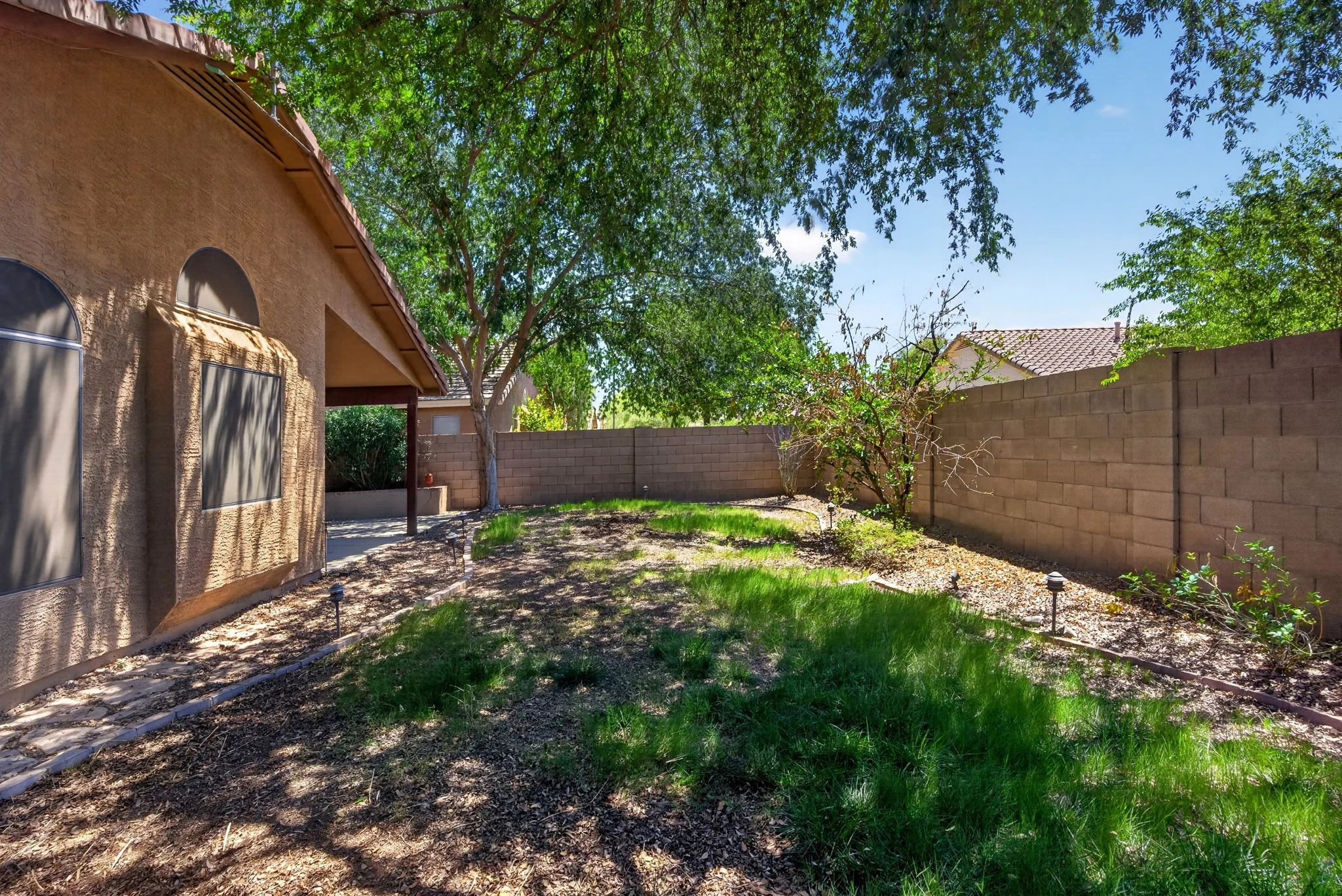 A backyard with a beige stucco house on the left, a brick wall fence on the right, tall green trees, a patchy lawn, and a blue sky.