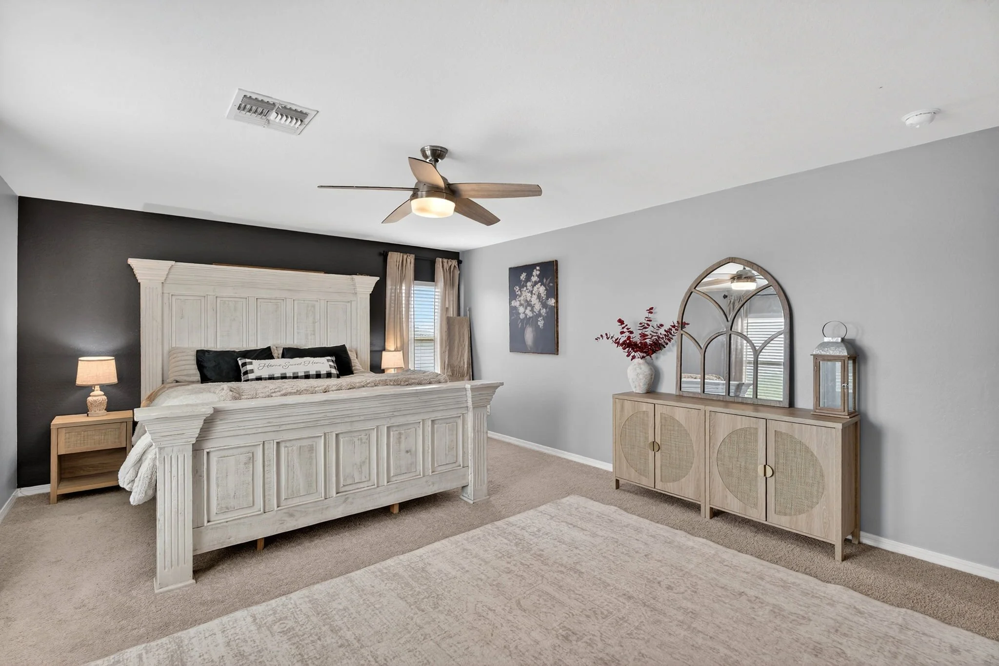 A bedroom with a white bed, nightstand, and lamps, a dark accent wall, light-colored curtains, a decorative mirror, and a dresser with a vase of red flowers.