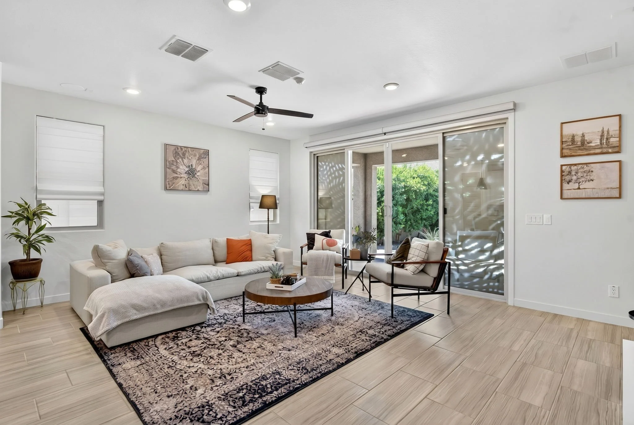 Living room with white couch, black armchair, coffee table, and artwork on the walls, large sliding glass door with outside view, and ceiling fan.