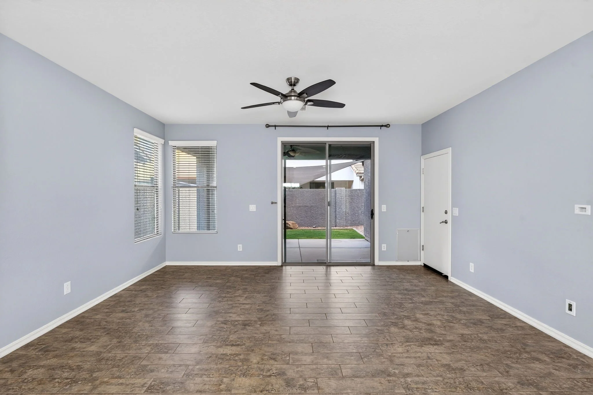Empty living room with light blue walls, wood flooring, large windows, a ceiling fan, and a sliding glass door leading outside.