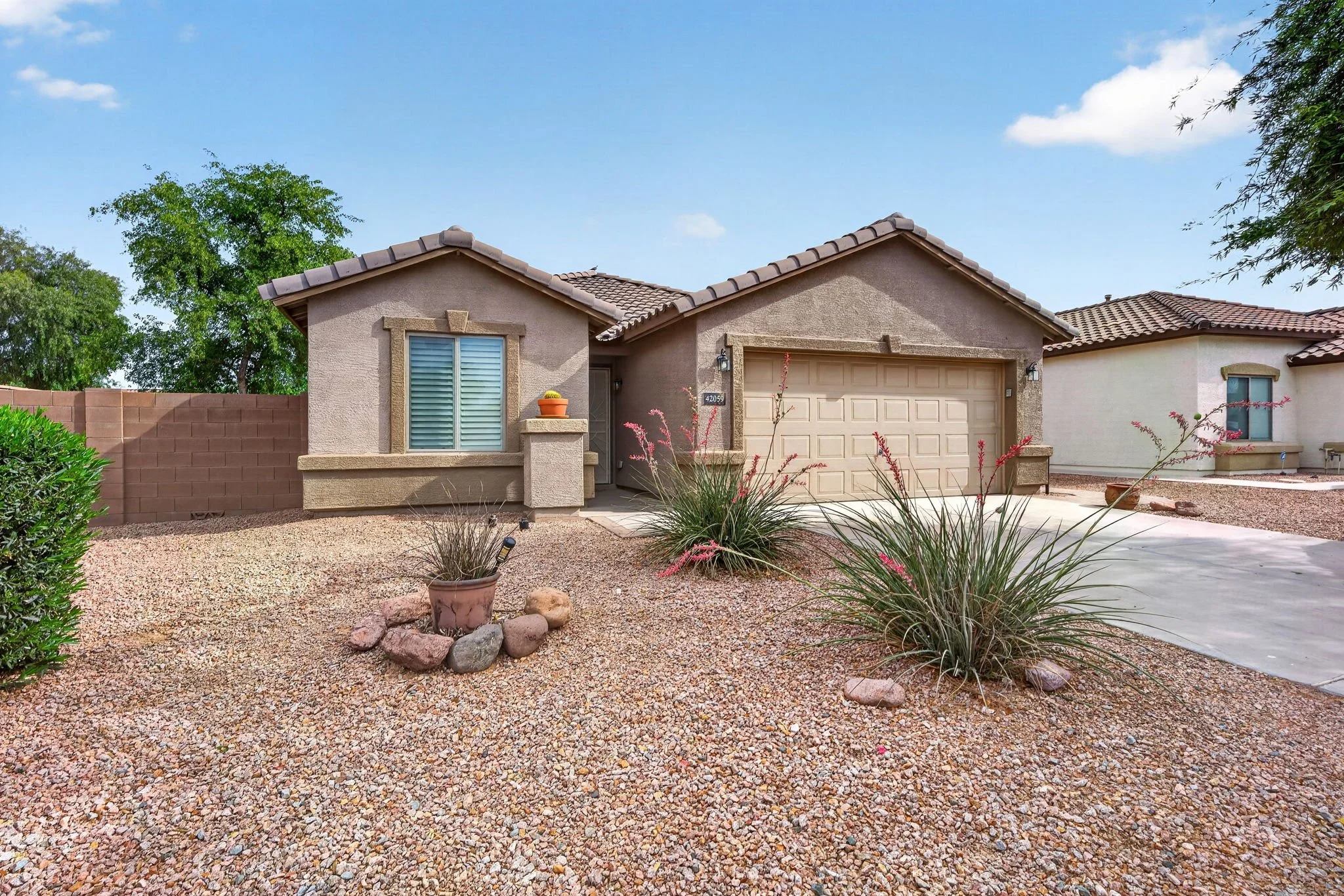 Front view of a beige stucco house with a brown tiled roof, a window with shutters, a garage door, and desert landscaping with rocks, plants, and a brick wall, under a partly cloudy sky.