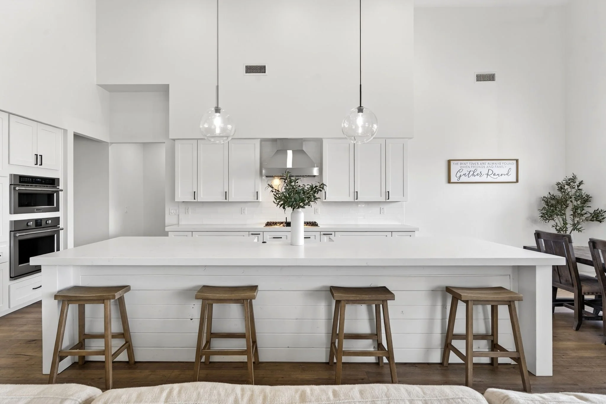 Modern, white kitchen with kitchen island, hanging glass pendant lights, and white cabinetry. Wooden stools at the island and framed wall art reading 'Gather Round' with indoor plants nearby.