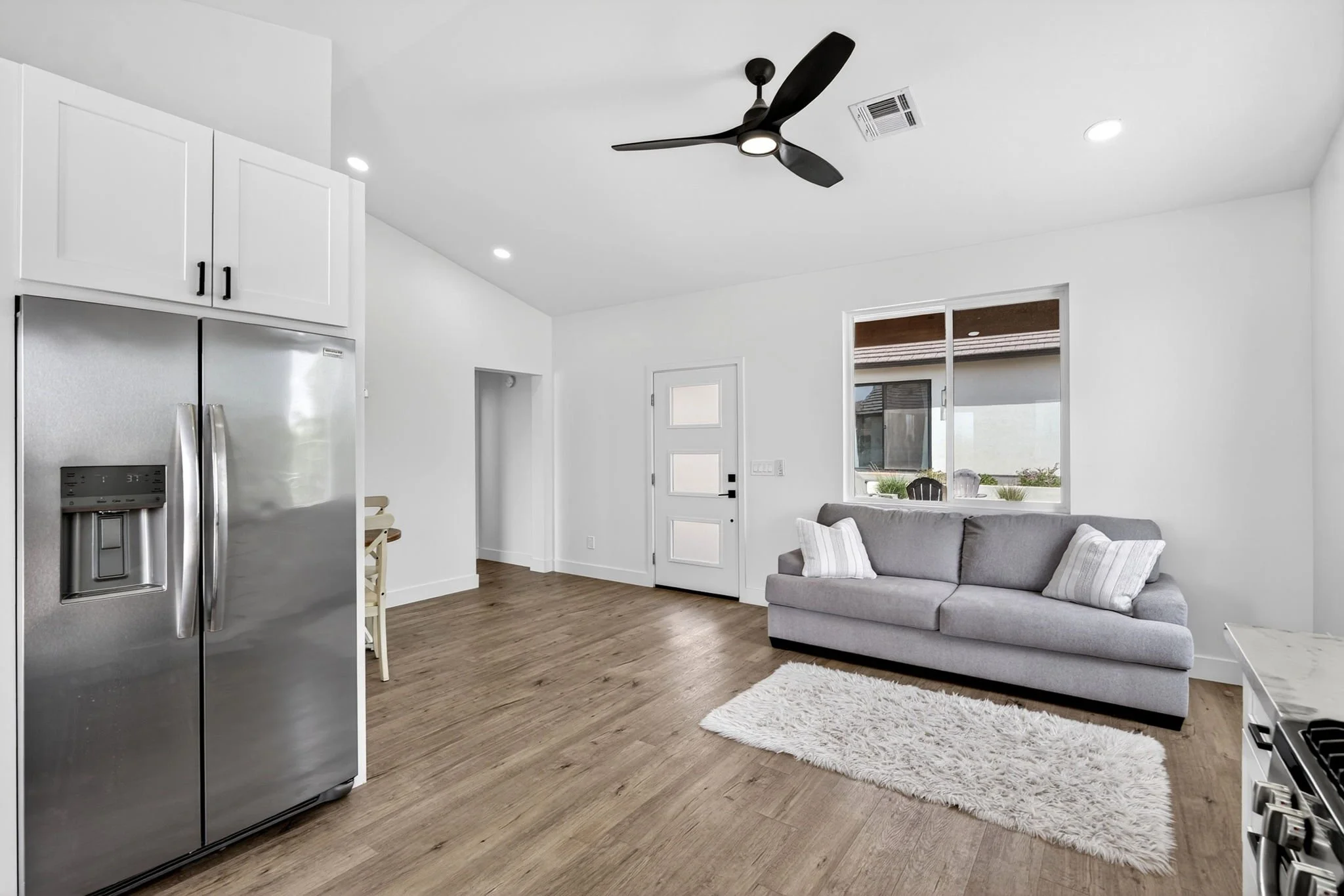 Modern living room with a gray sofa, white pillows, a white shag rug, wooden flooring, a window with a view outside, a white door, and a ceiling fan with black blades.