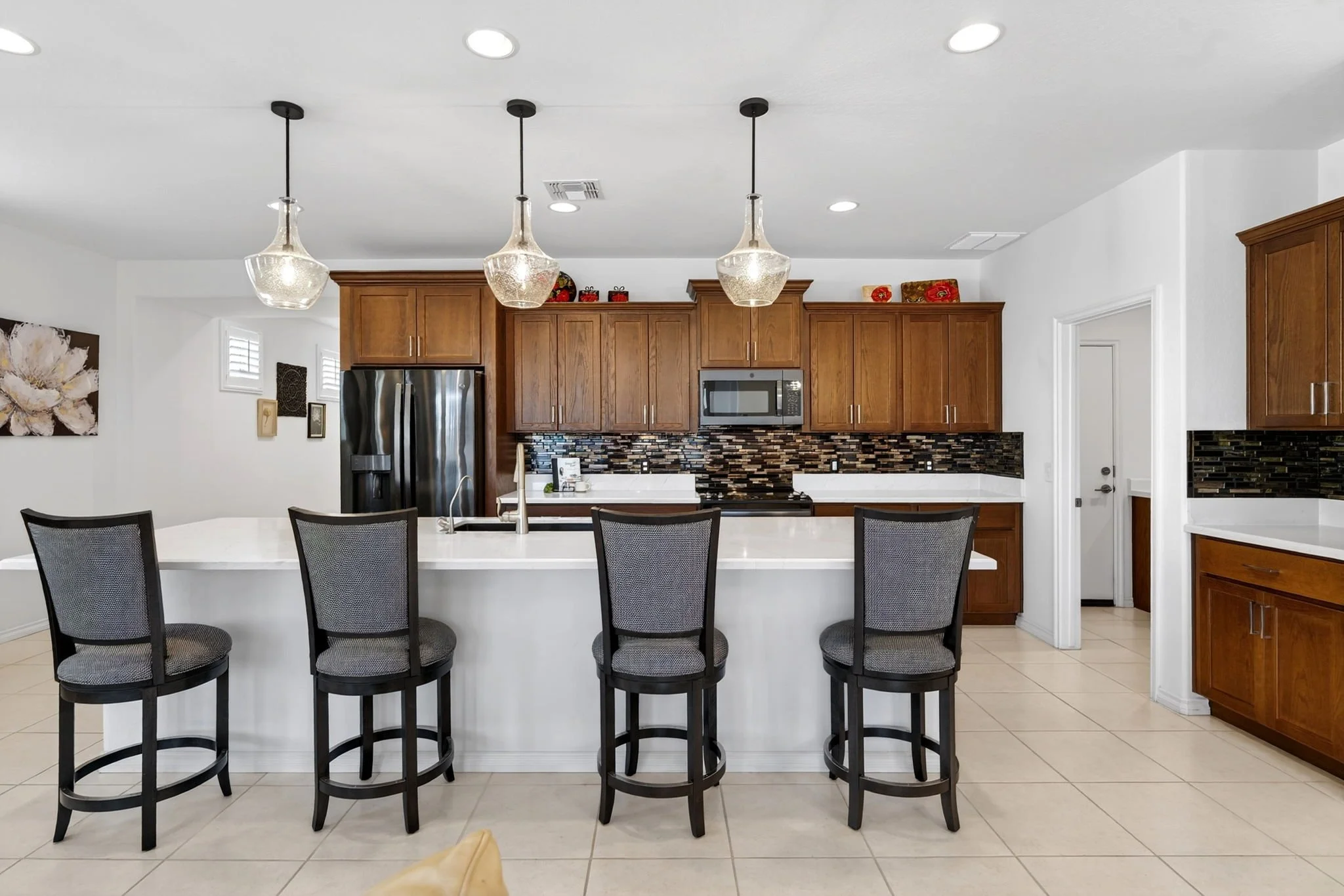 Modern kitchen with white countertops, wooden cabinets, black and gray backsplash, and three pendant lights hanging over an island with black chairs.