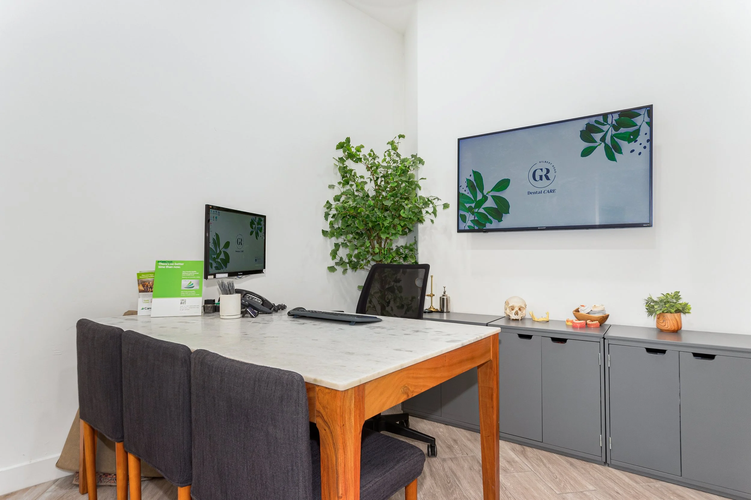 Office reception area with a white wall, a grey cabinet, a desk with a marble top, a computer monitor, a desk chair, and decorative items including a skull model and plants. A large screen mounted on the wall displays a logo for "GR Dental CARE."