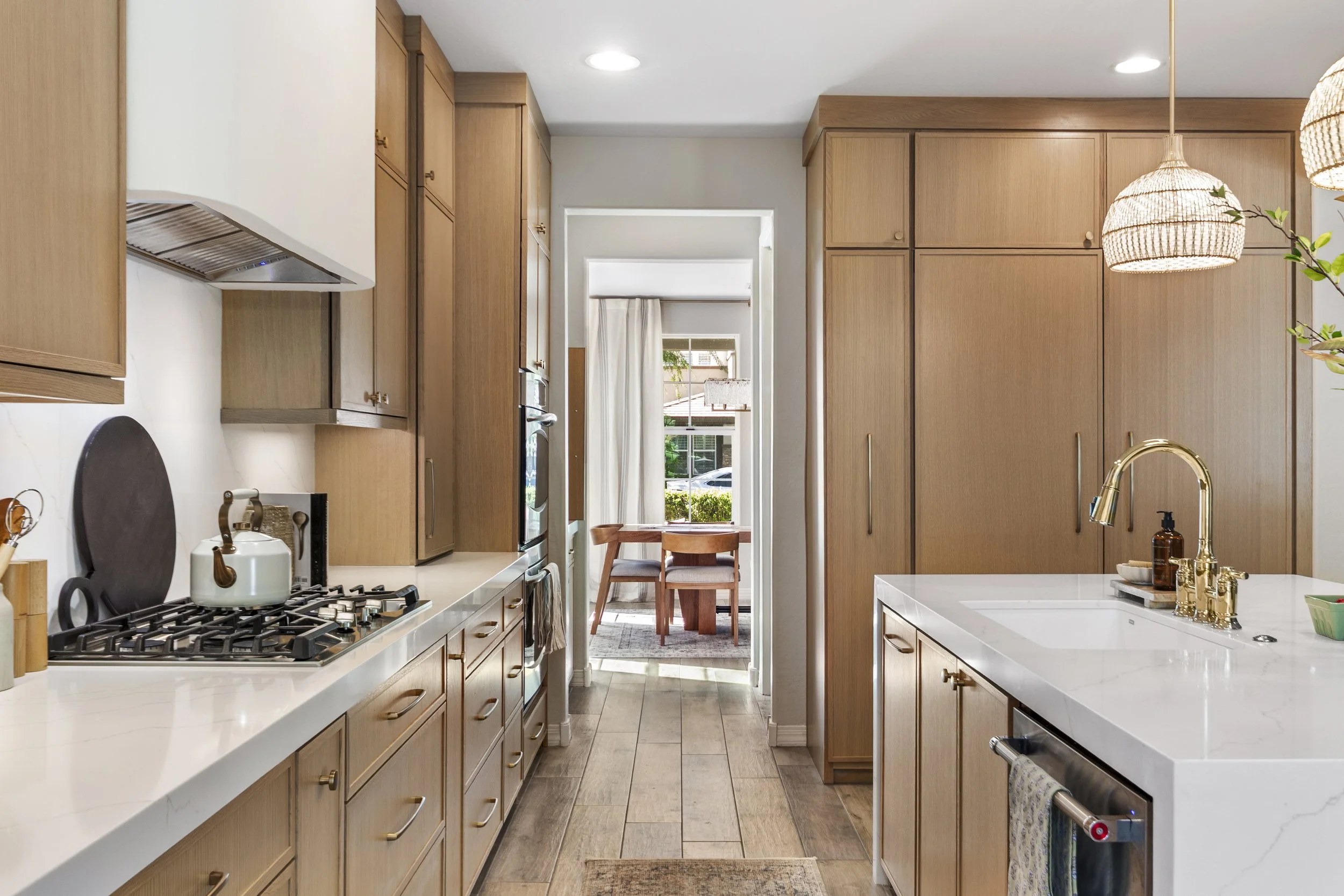 A modern kitchen with light wooden cabinets, white countertops, and stainless steel appliances. Sunlight streams in from a window at the back, and a small dining table with chairs is visible through an open doorway.