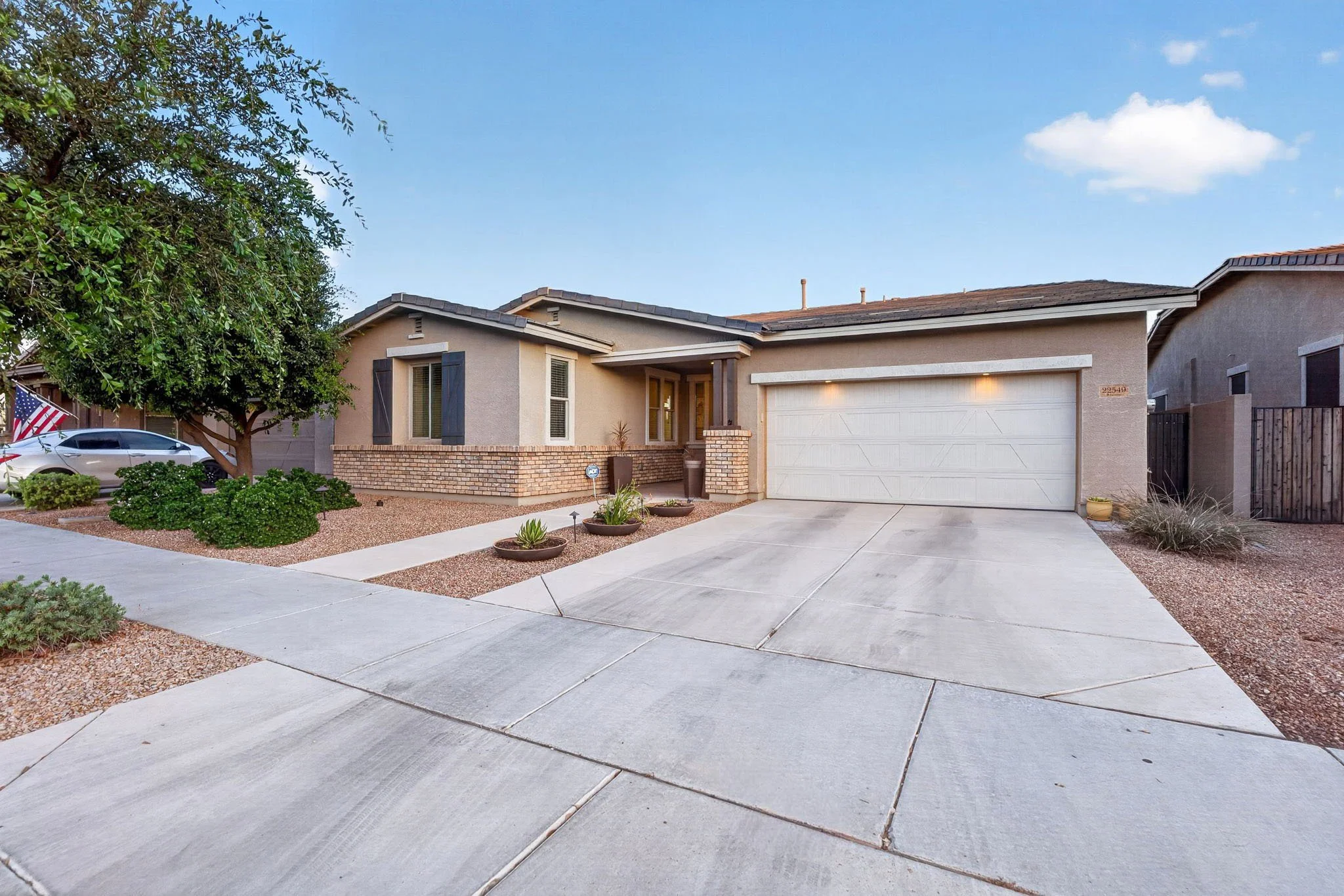 Front view of a single-story house with a light-colored stucco exterior, black shutters, a brick base, a large garage door, and a small porch with potted plants. The driveway is concrete, surrounded by landscaped yard with bushes and gravel. There is