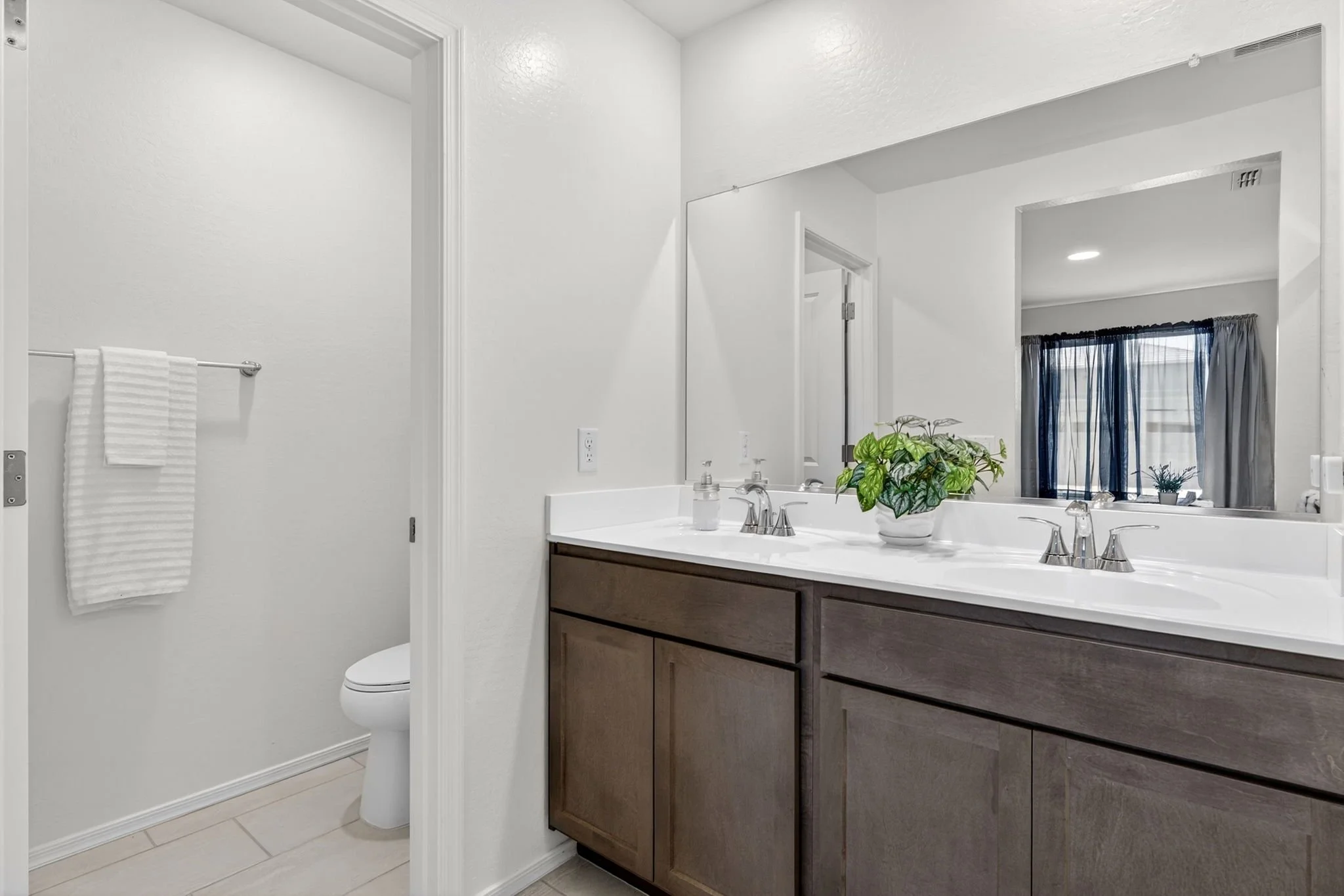 Bathroom with double sink vanity, large mirror, and a plant on the counter. Visible part of a toilet and a towel hanging on a rod. Window with curtains in the background.