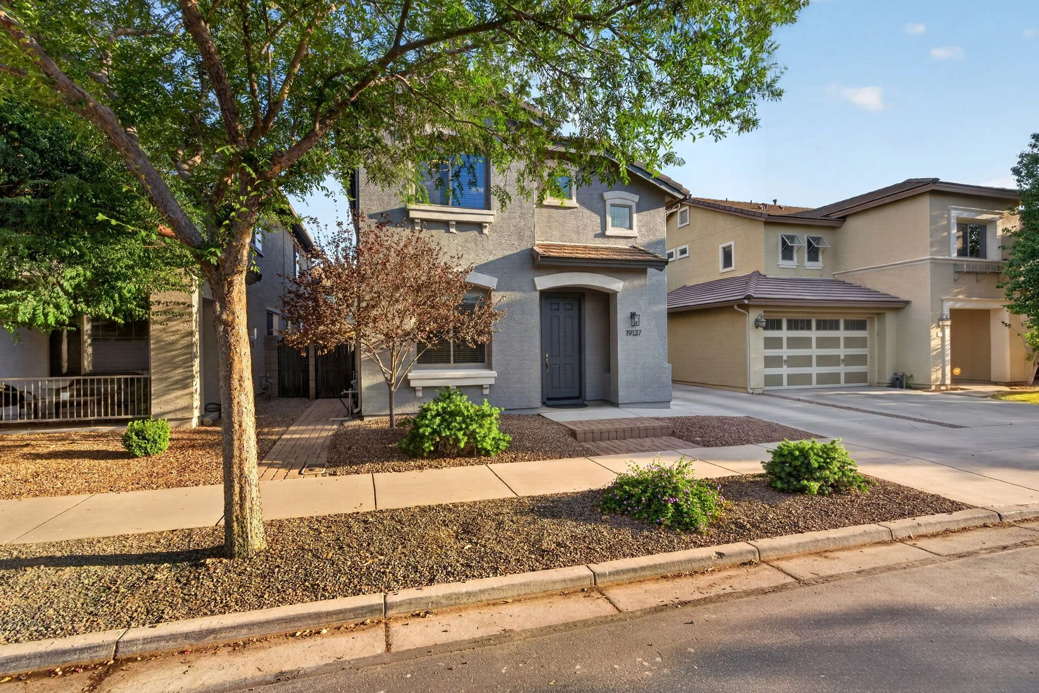 A suburban street with a gray two-story house, a driveway, and trees and bushes in the front yard.