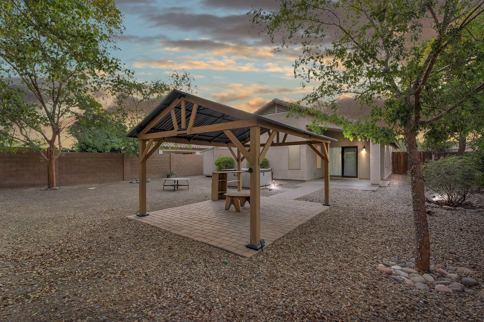 Backyard patio with a small covered wooden pergola, trees, gravel ground, a brick wall, and a house with a sliding glass door at sunset.