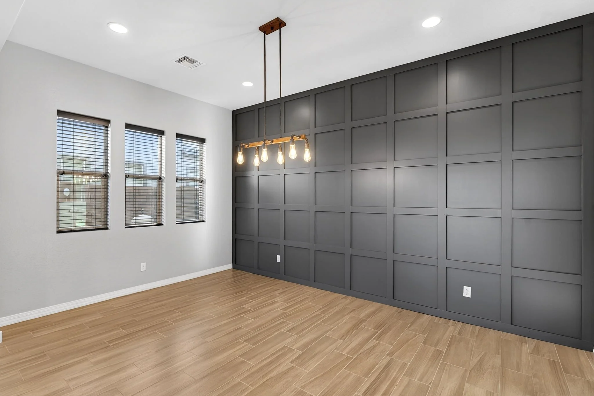 Interior of a modern room with a gray accent wall featuring paneled design, three windows with blinds, and a wooden floor. A contemporary chandelier with exposed bulbs hangs from the ceiling.
