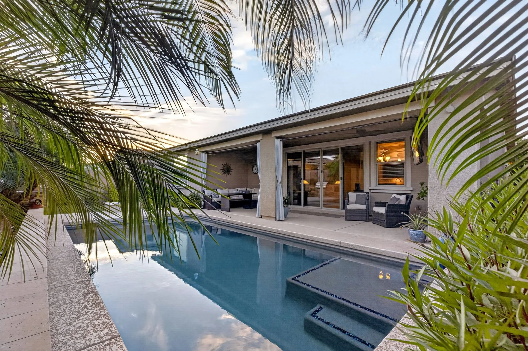A backyard swimming pool area with outdoor furniture, surrounded by tropical plants and palm trees, at sunset.