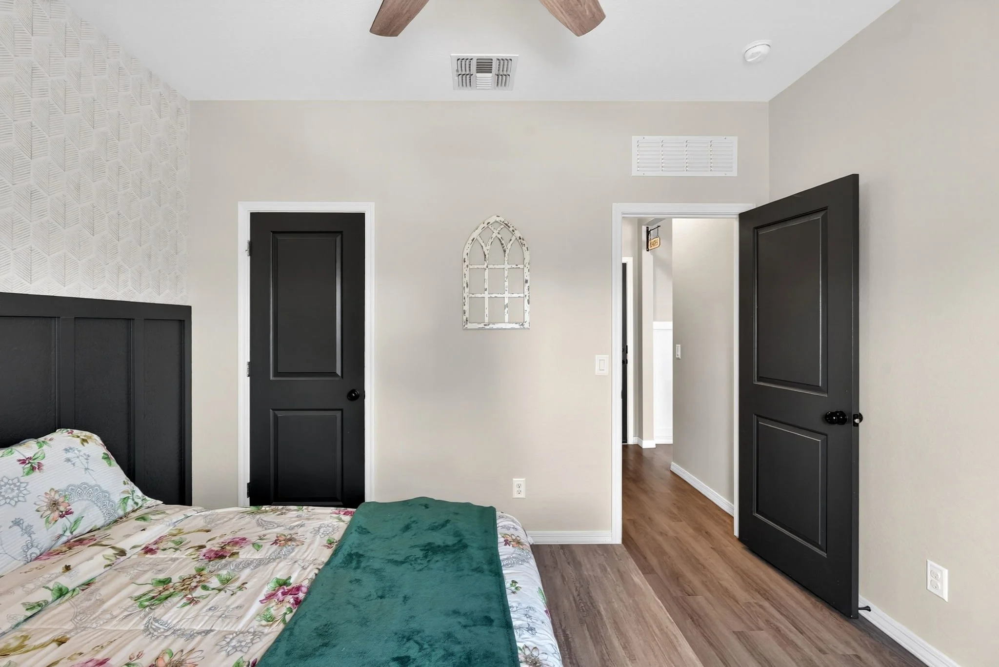 A bedroom with a bed featuring a floral quilt and pillows, black doors and trim, a decorative window on the wall, wood flooring, and a ceiling fan.