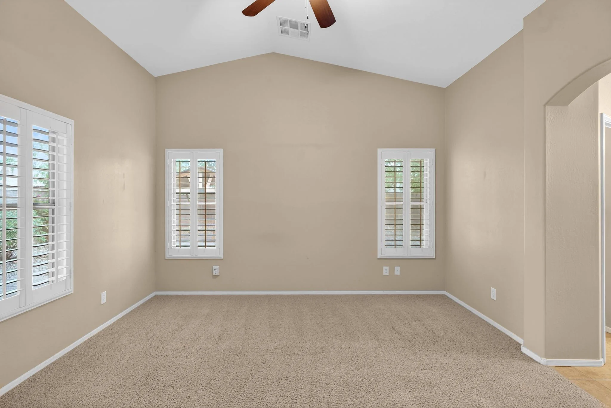 Empty beige living room with three windows having white shutters, a ceiling fan, and beige carpet.