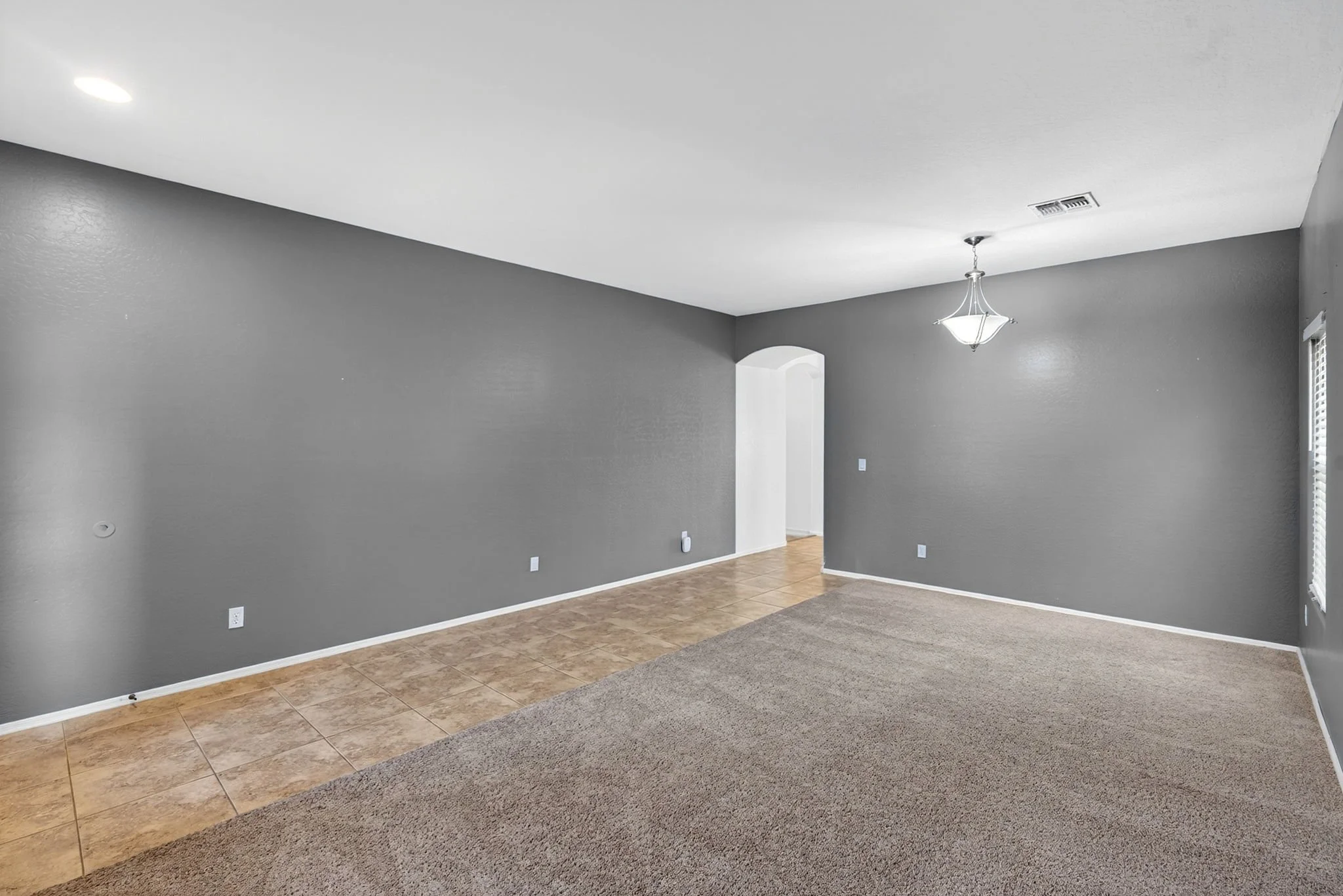 Empty living room with gray walls, beige tiled and carpeted flooring, a modern hanging ceiling light, and a window with white blinds.