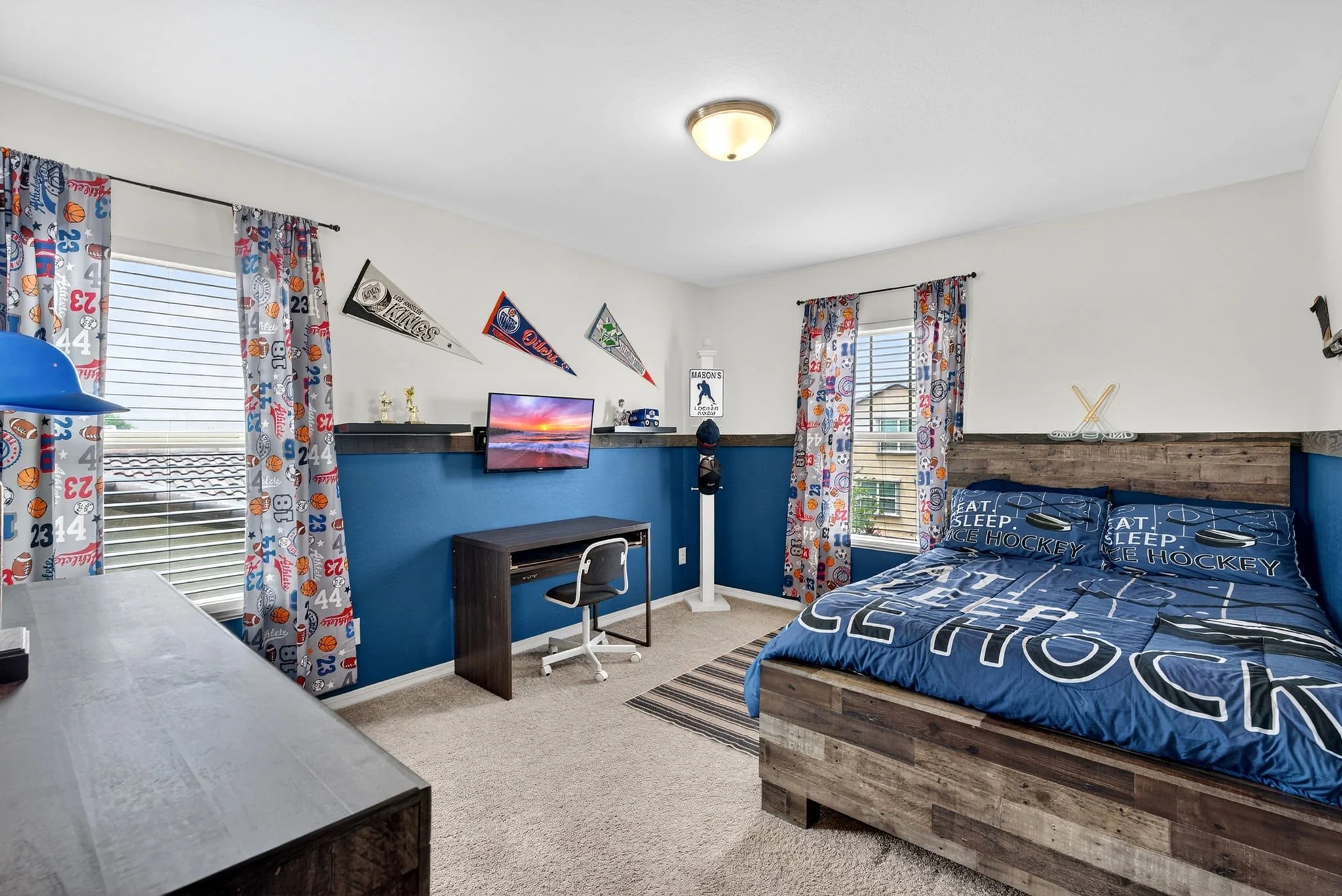 A bedroom with two windows, sports-themed curtains, a bed with a hockey-themed bedspread, a small desk with a chair, a wall-mounted TV, and sports pennants hanging on the wall.