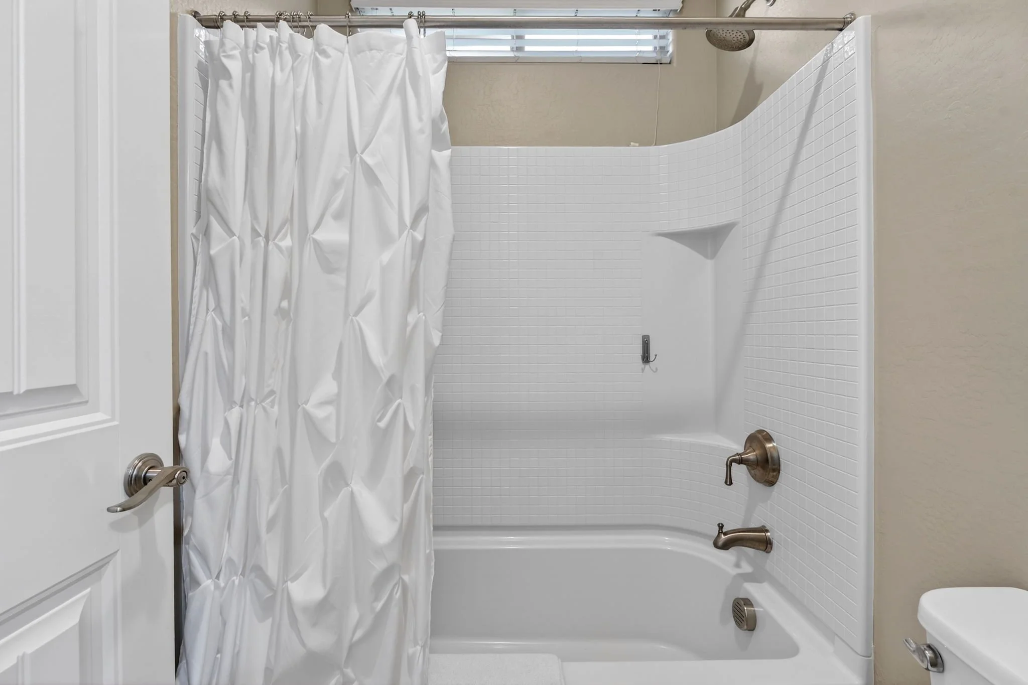 Empty bathtub with white tiled walls and a shower curtain on a metal rod inside a bathroom. A small window with blinds above the shower. Silver faucet and knobs on the side of the bathtub.