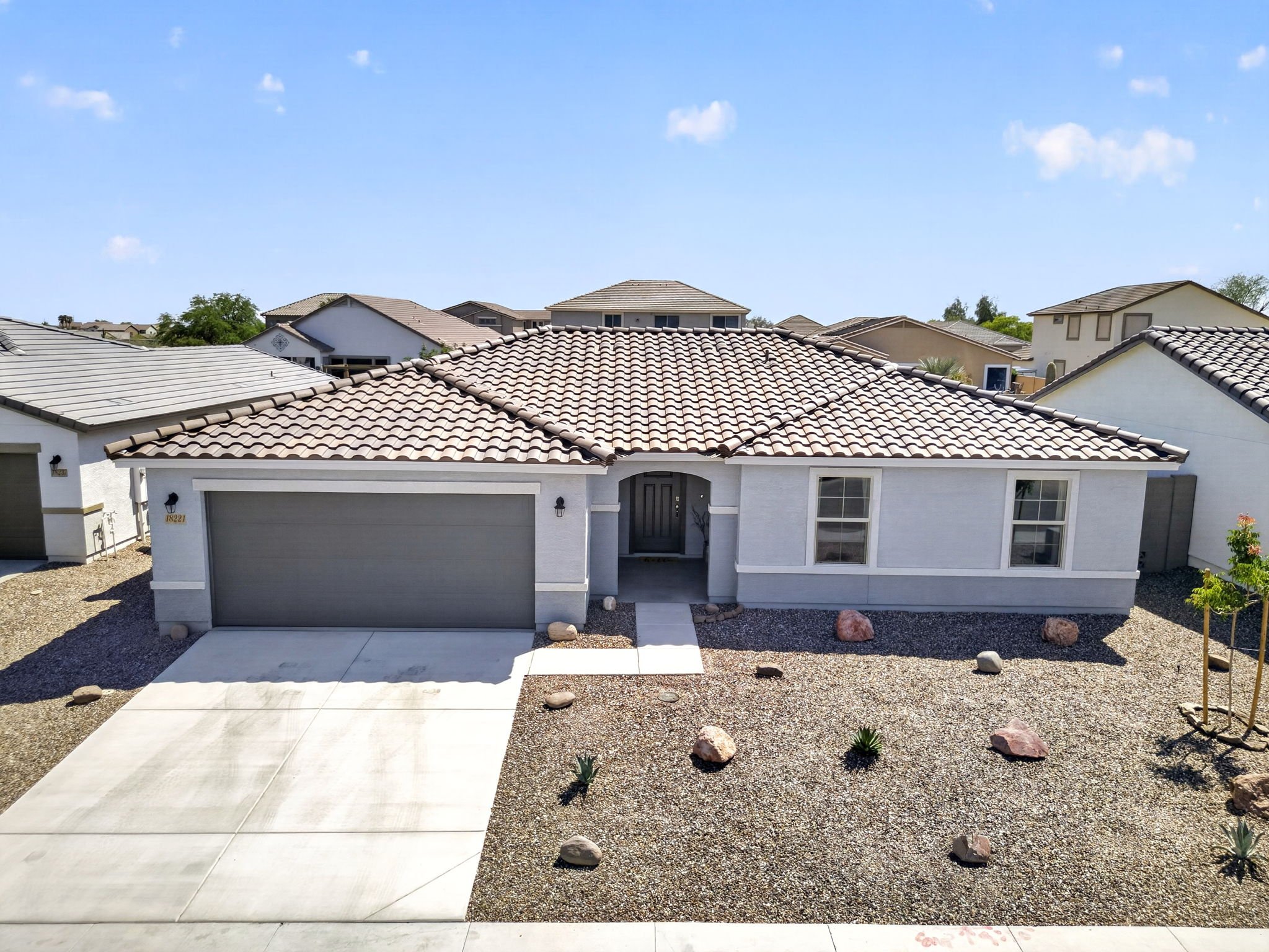 Front view of a modern suburban house with a tiled roof, white exterior, and a driveway leading to a garage. The front yard features rocks, desert plants, and small bushes, with rocks placed along the landscape. The sky is clear with a few clouds.