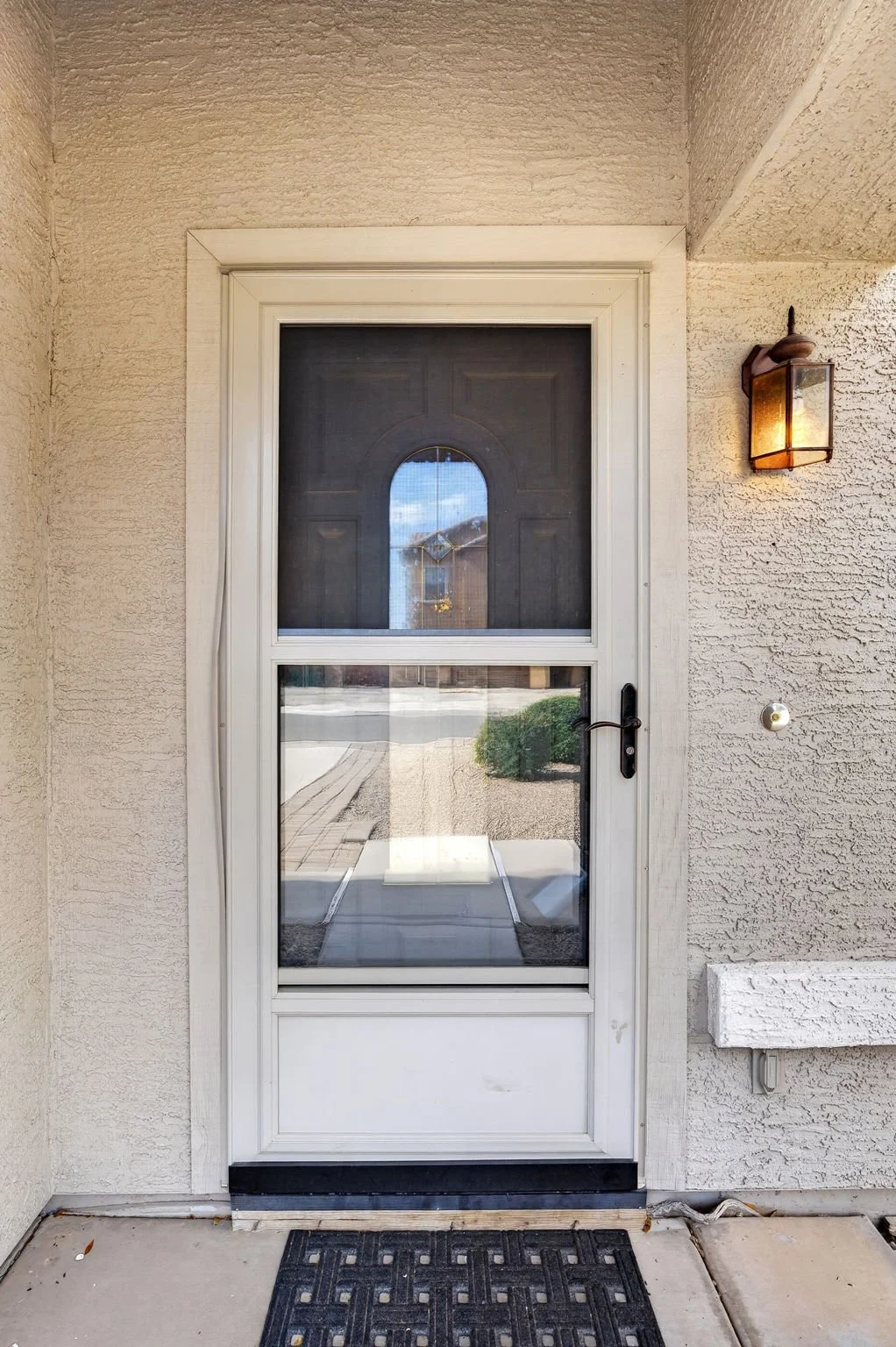 Front door with a screen door and black handle, beige textured exterior wall, wall-mounted lantern light to the right of the door, small white fixture to the right of the door, black doormat on concrete porch.