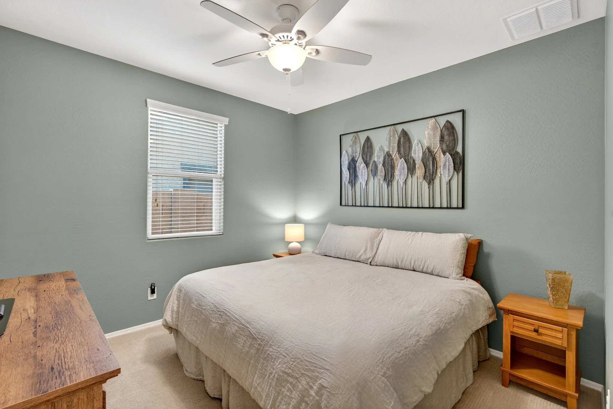 A bedroom with light green walls, a window with blinds, a ceiling fan with light, a framed leaf artwork above the bed, a beige bedspread and pillows, a small wooden nightstand with a lamp, a wooden dresser, and a decorative glass object on the nights