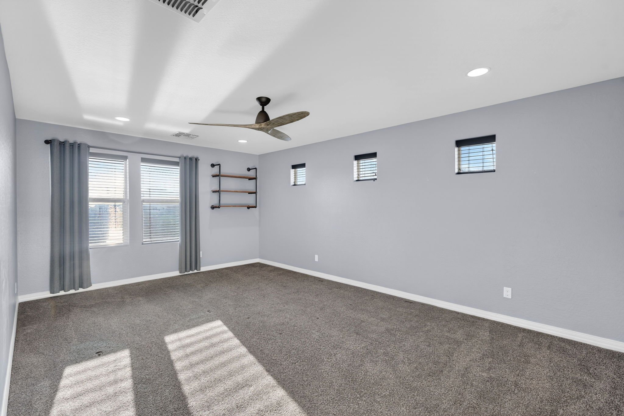 Empty room with gray walls, dark carpet, multiple small windows with black blinds, ceiling fan, and built-in shelves.