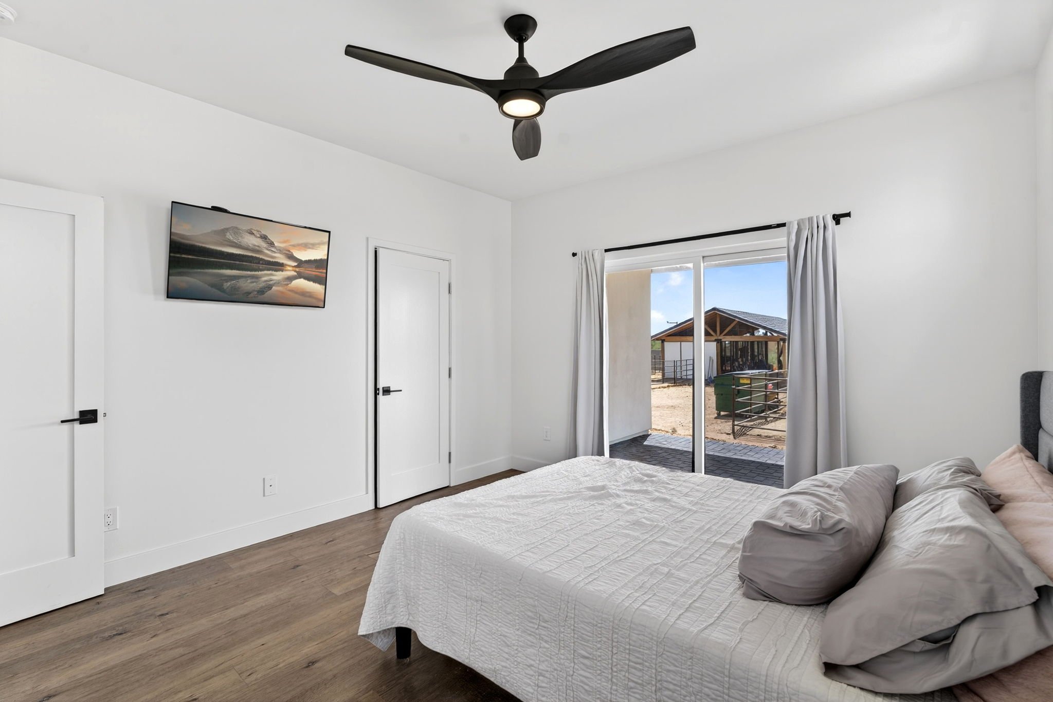 Bedroom with white walls, a bed with white bedding and pillows, a ceiling fan, a wall-mounted TV, and sliding glass doors with white curtains leading outside to a patio.
