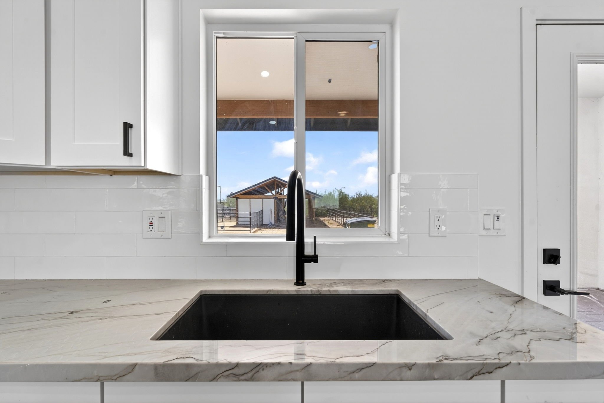 View of a kitchen with a white marble countertop, black sink, white cabinets, and a large window showing an outdoor scene with a barn and blue sky.