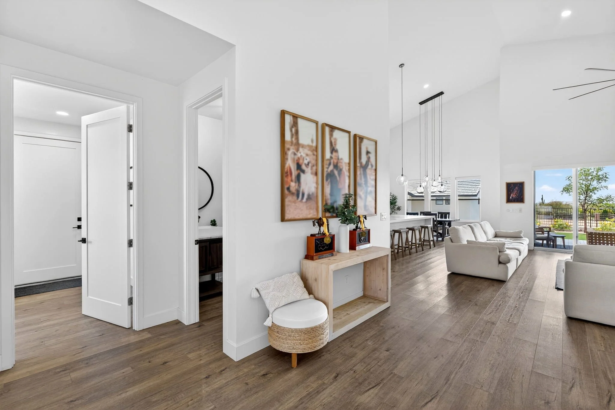Open-concept living room and kitchen with white walls, wooden floors, and large windows showing an outdoor yard. Decor includes framed photos, a white sofa, and a small chair with a woven basket at its side.