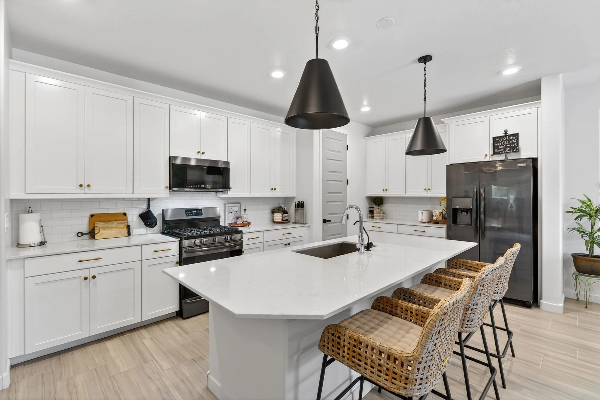 Modern white kitchen with island, black hanging pendant lights, stainless steel appliances, and wicker bar stools.