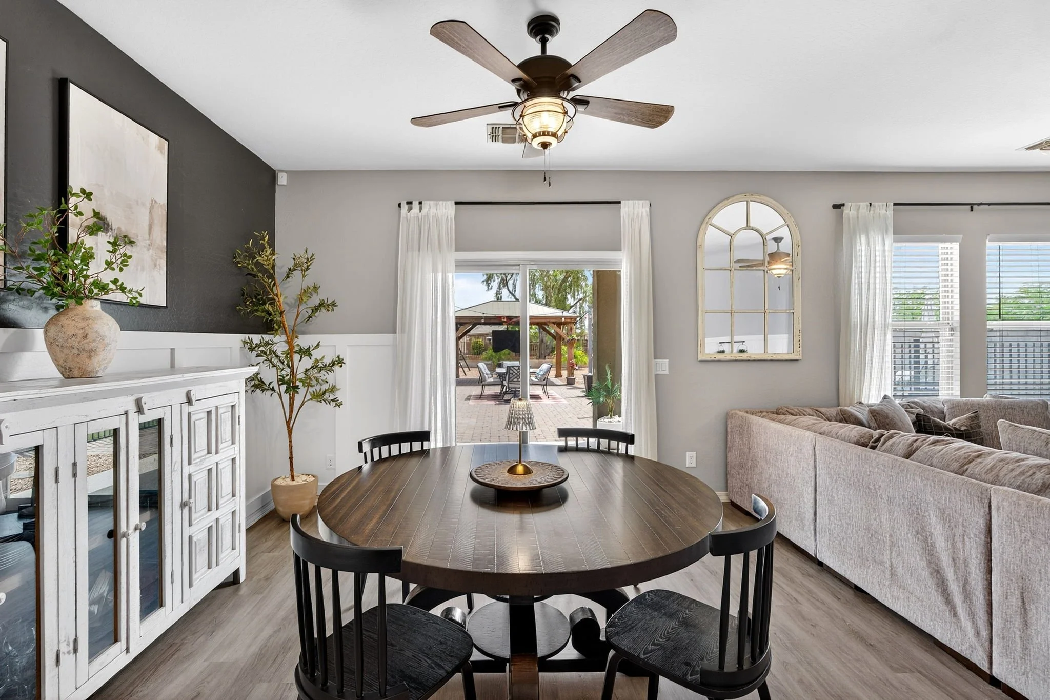 Interior of a living room with a round wooden dining table surrounded by black chairs, a beige sectional sofa, white wainscoting, a ceiling fan with wooden blades, large windows with white curtains, a sliding glass door leading outside, and decorativ