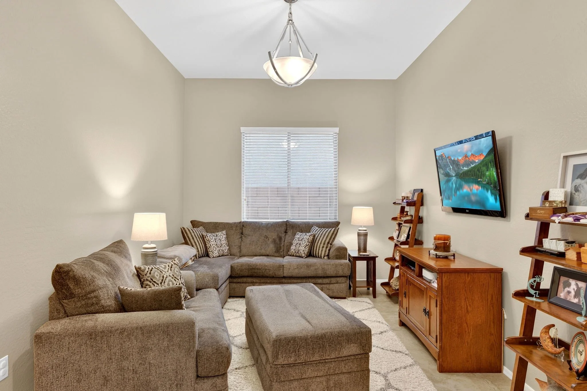 Living room with beige walls, a large sectional sofa with throw pillows, a wooden entertainment center, a flat-screen TV, and a window with white blinds. There are also lamps and personal photos on the furniture.