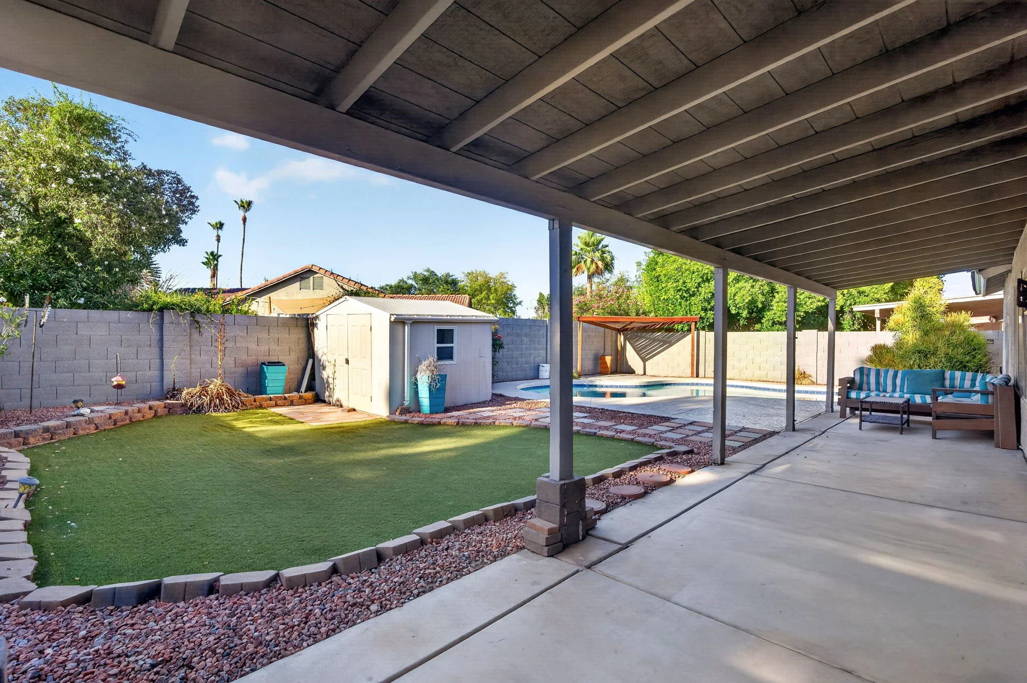 Backyard with a covered patio, a swimming pool, a small garden bed, a shed, and remote landscaping surrounded by a cinder block fence, with trees and neighboring houses in the background.
