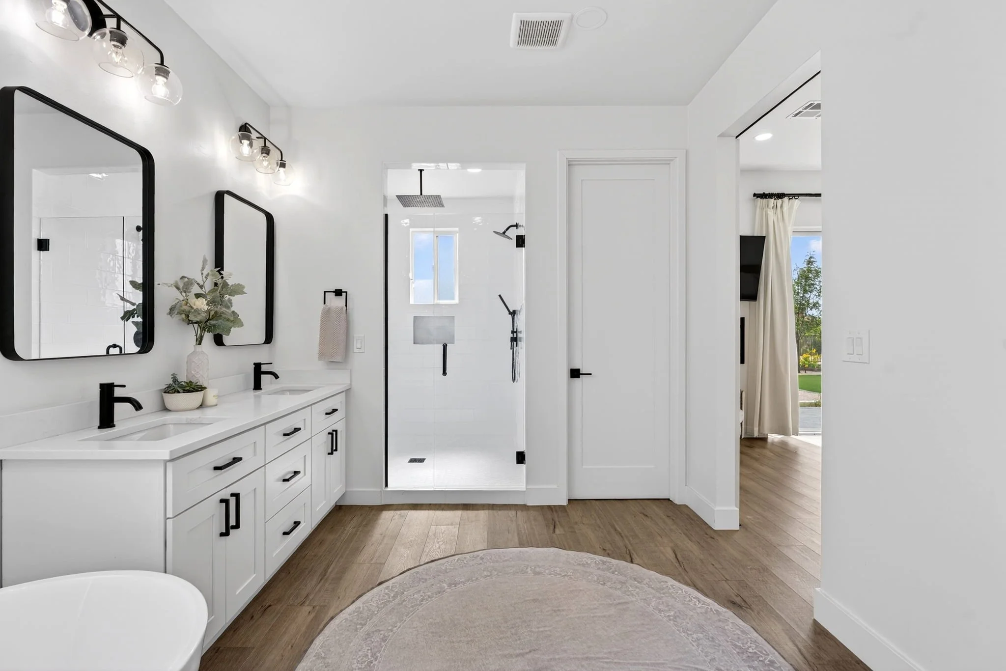 Modern bathroom with white vanity, black fixtures, mirrors, and a glass enclosed shower with a rainfall showerhead, wooden floors, and a window showing a blue sky and green trees.