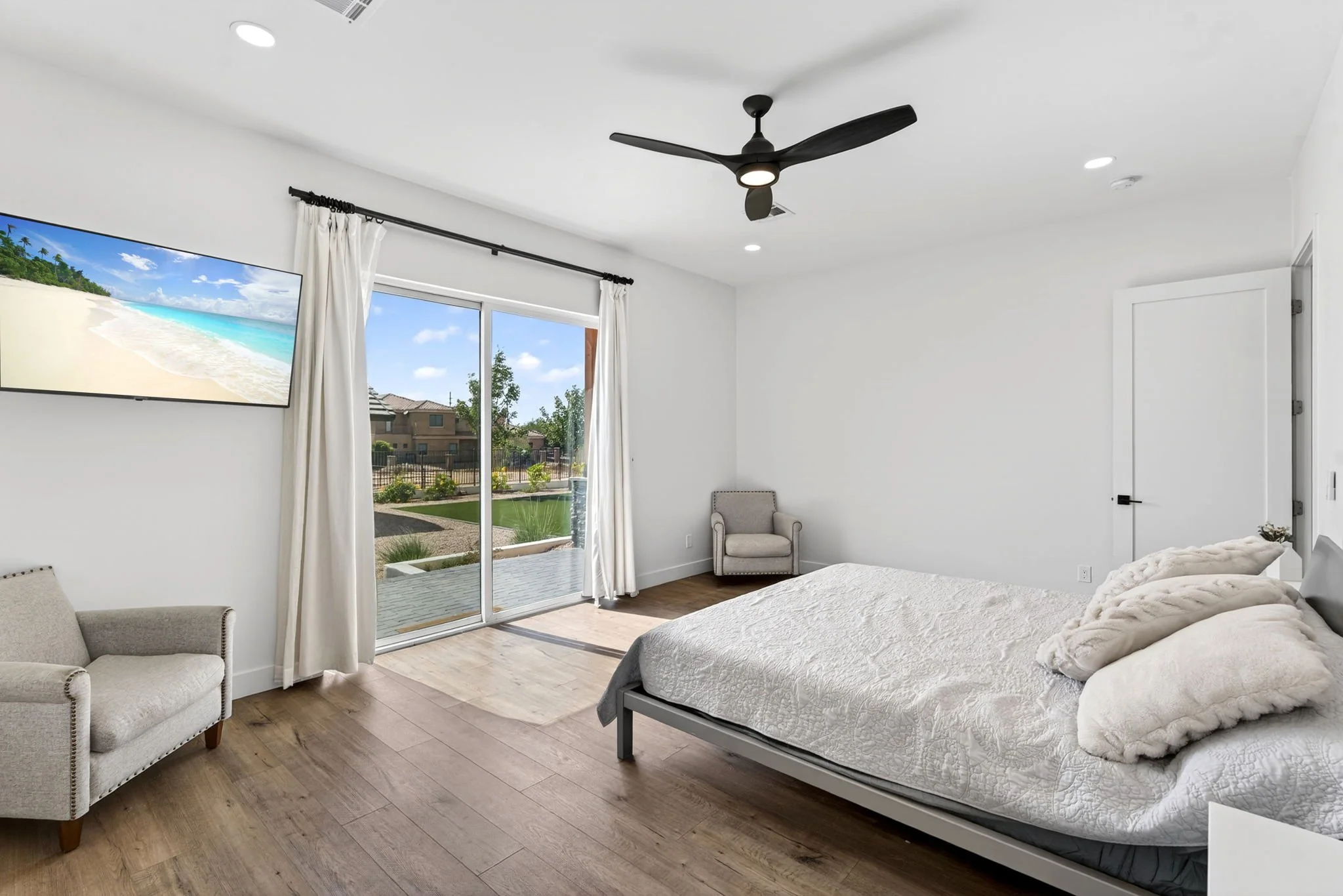 A bedroom with white walls, hardwood floors, a large window with white curtains, a mounted flat-screen tv showing a beach scene, a ceiling fan, a bed with white bedding and a textured throw blanket, a beige armchair, and a sliding glass door leading 