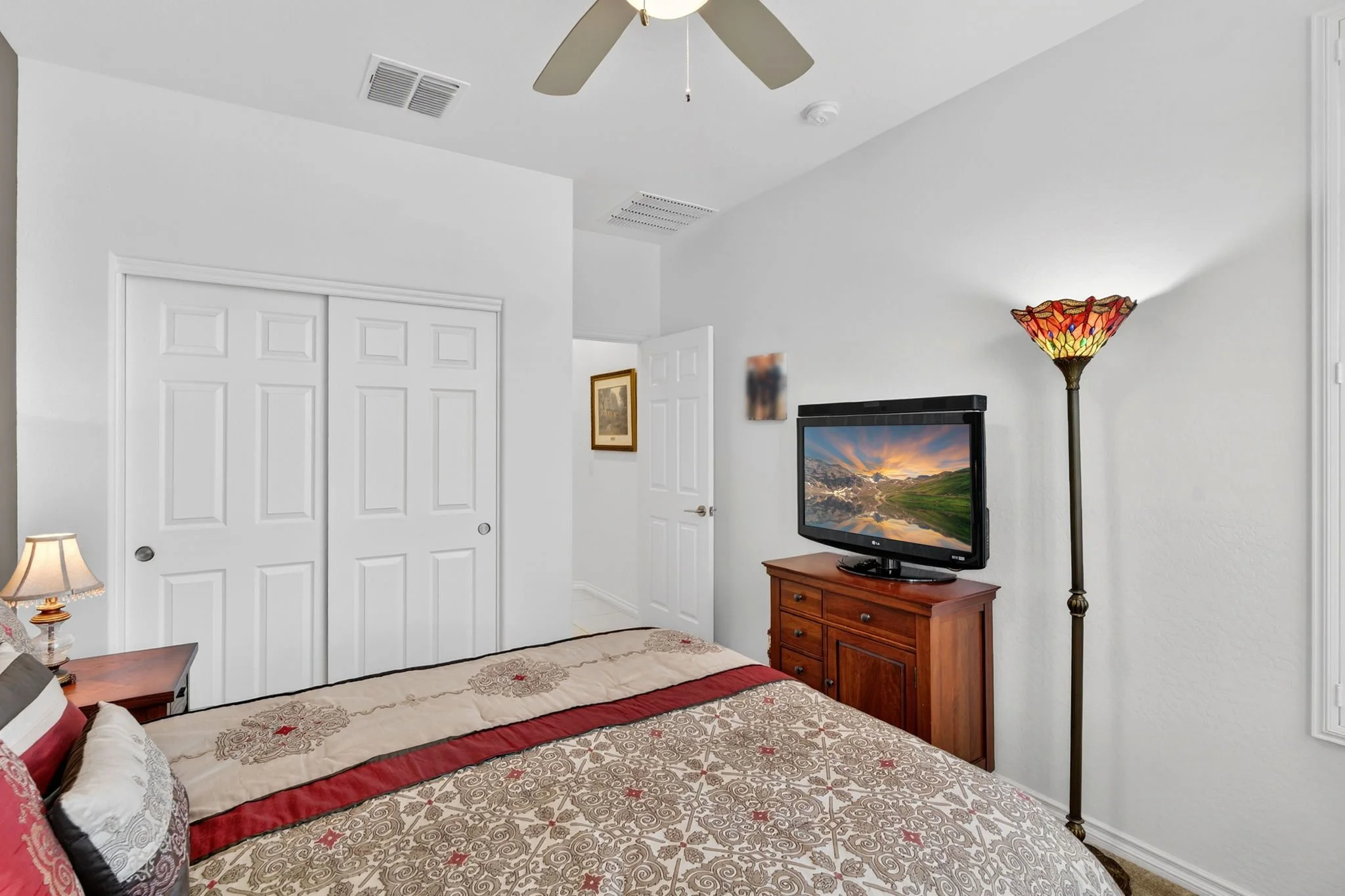 A bedroom with a bed, a wooden dresser with a television, a colorful stained glass floor lamp, and a ceiling fan. White walls and closet doors are visible.